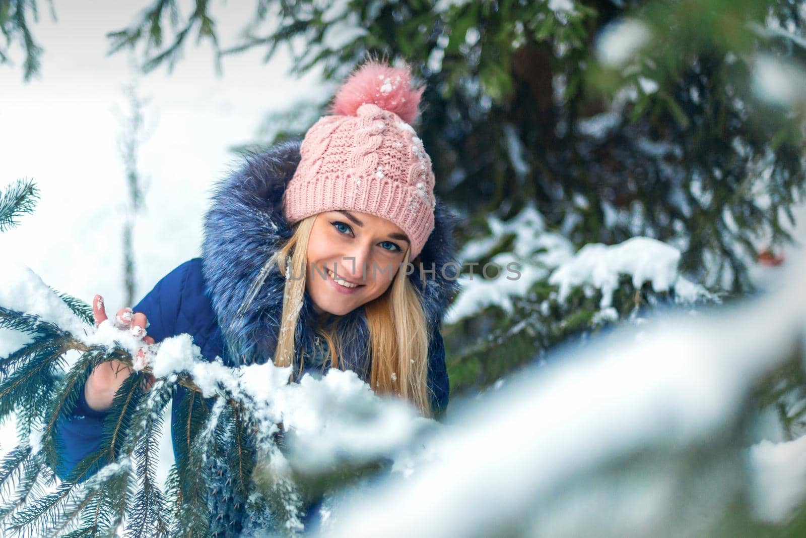 Portrait of beautiful smiling caucasian young woman standing in winter forest wearing blue jacket and pink hat