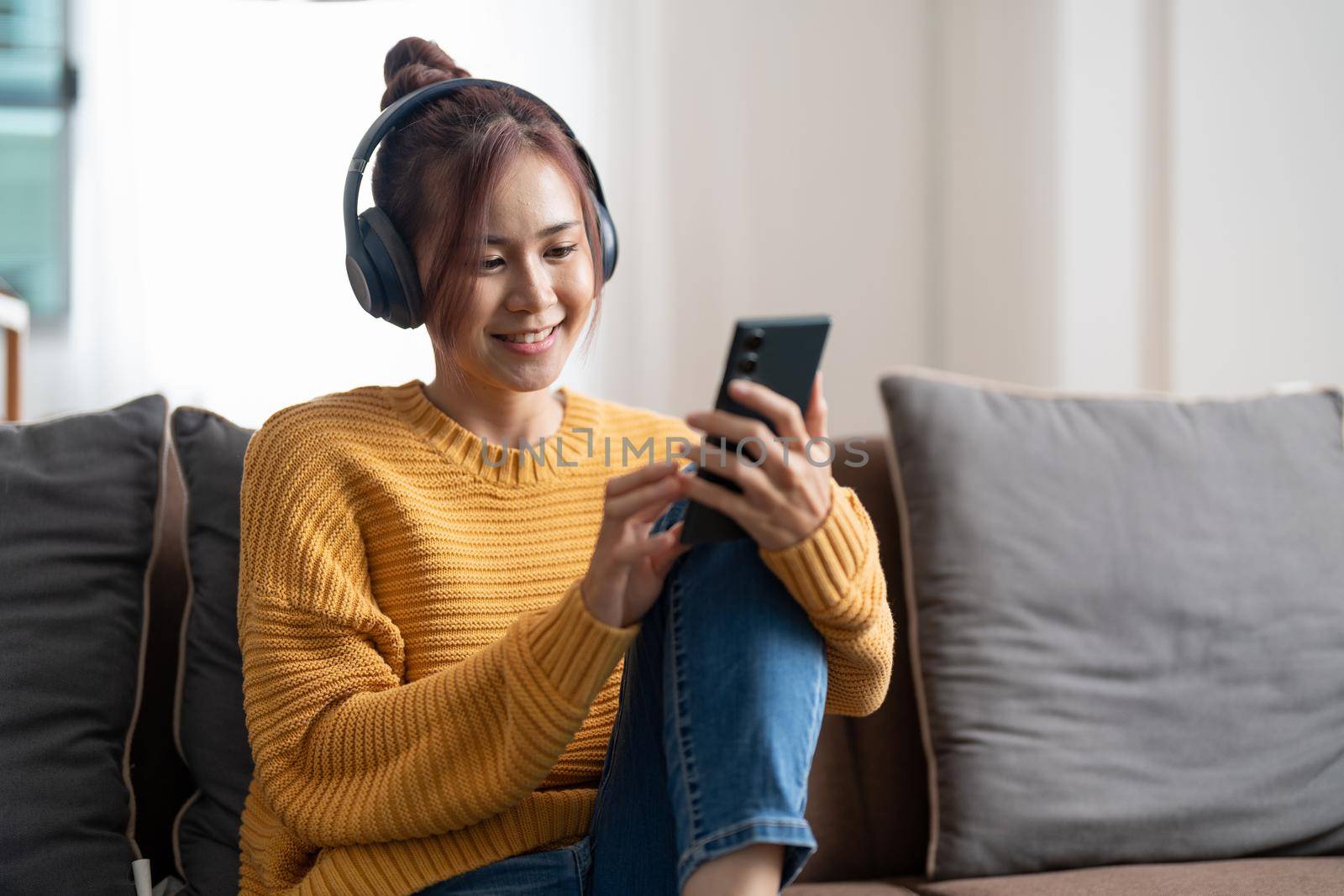 Freelance asian woman with mobile phone listening music in headphones and relax at home. Happy girl sitting on couch in living room. by nateemee