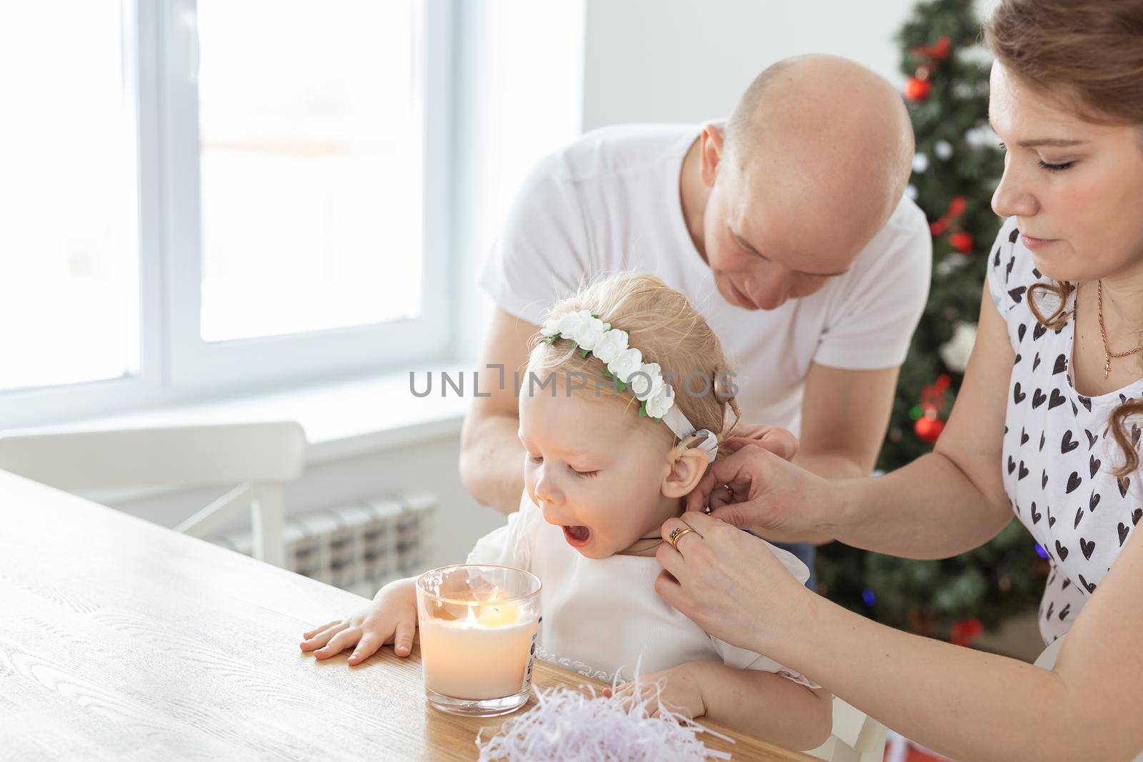 Mother and father helps to put on cochlear implant for their deaf baby daughter in christmas living room. Hearing aid and innovating medical technologies treatment deafness