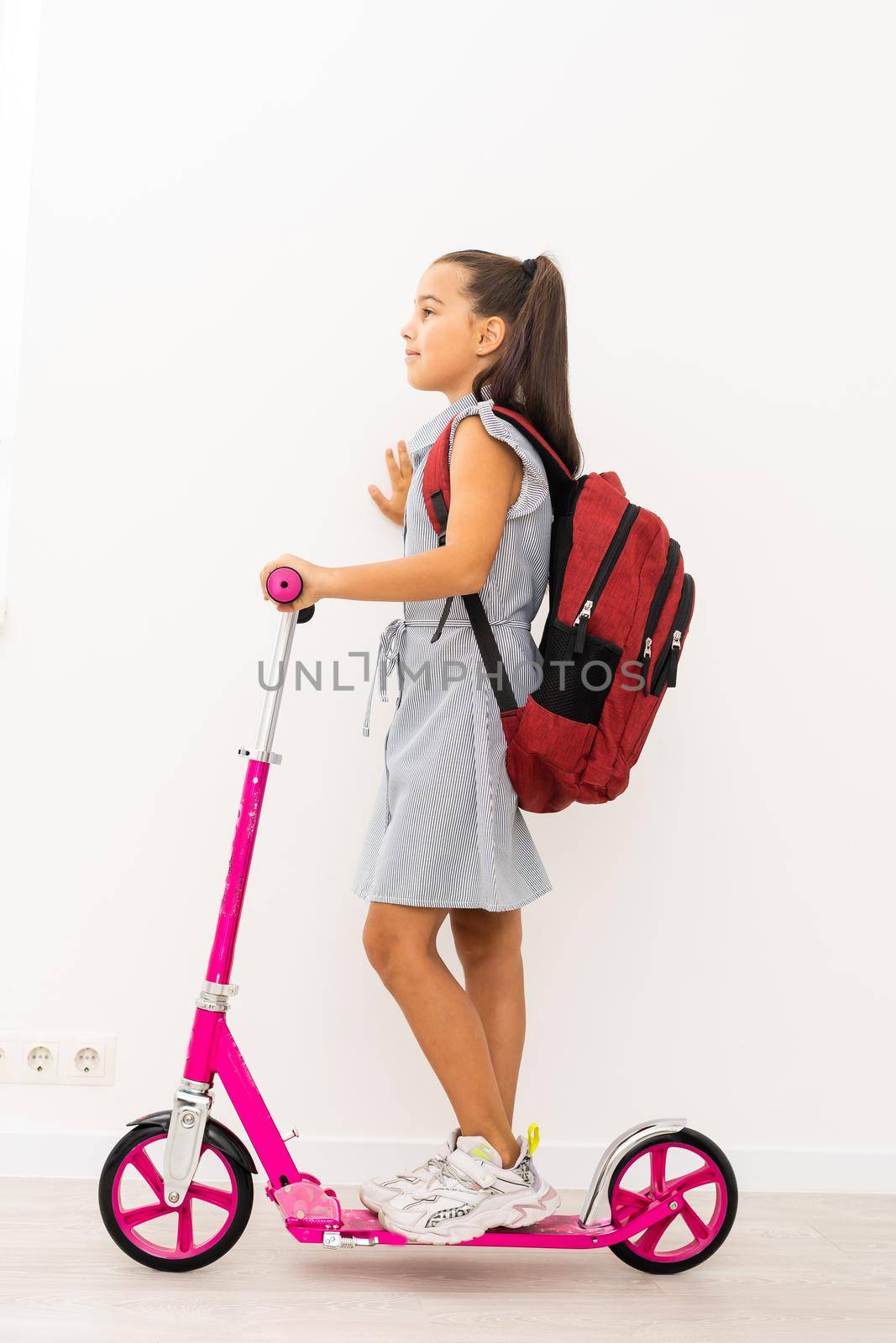 Full length profile shot of a schoolgirl with a backpack riding a scooter isolated on white background by Andelov13