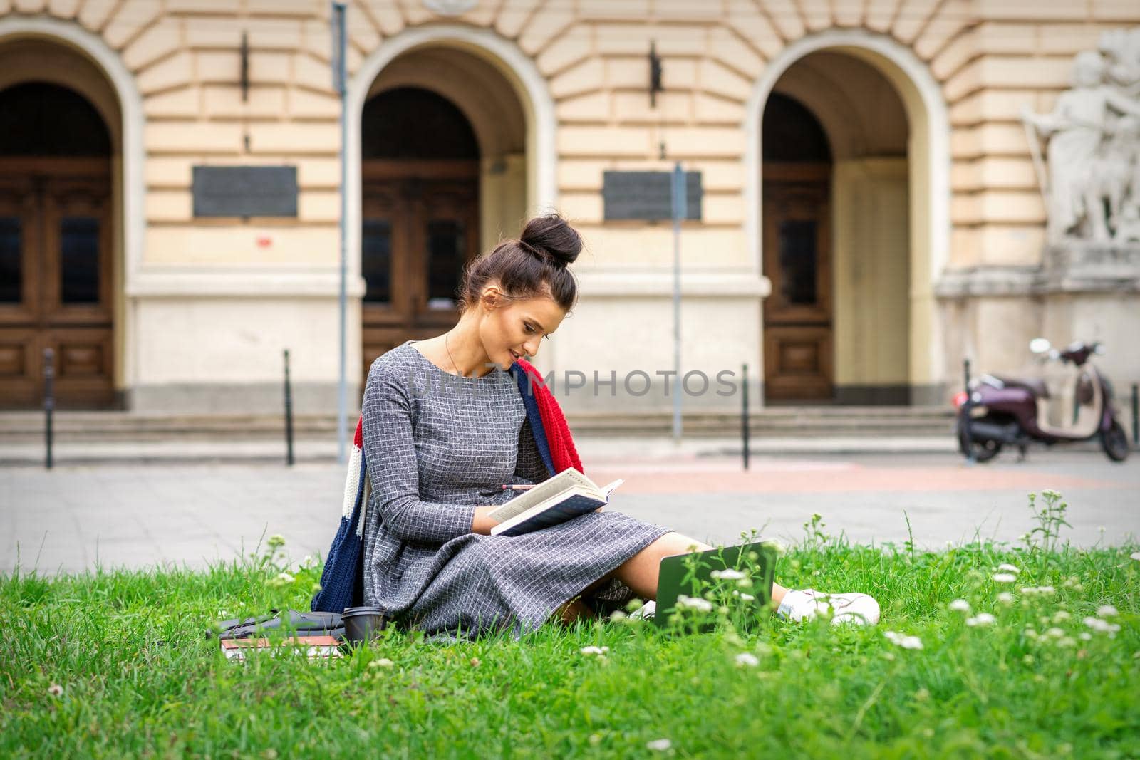 Young caucasian student woman reading the book sitting on the grass near university on the street