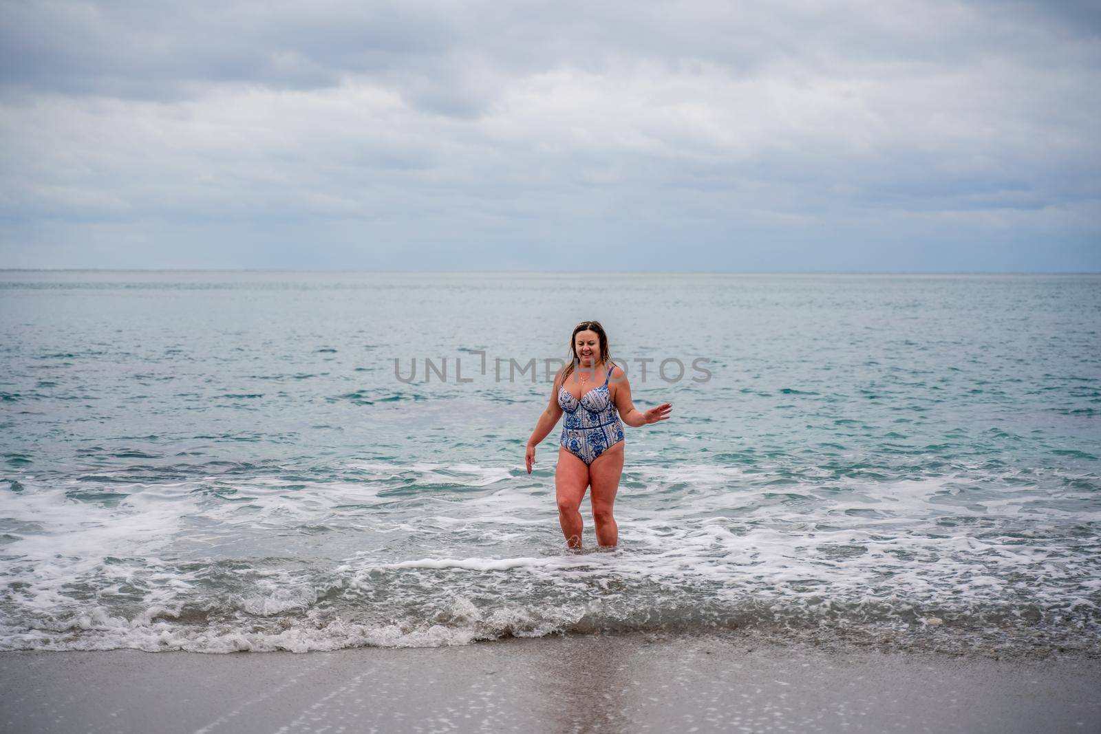 A plump woman in a bathing suit enters the water during the surf. Alone on the beach, Gray sky in the clouds, swimming in winter