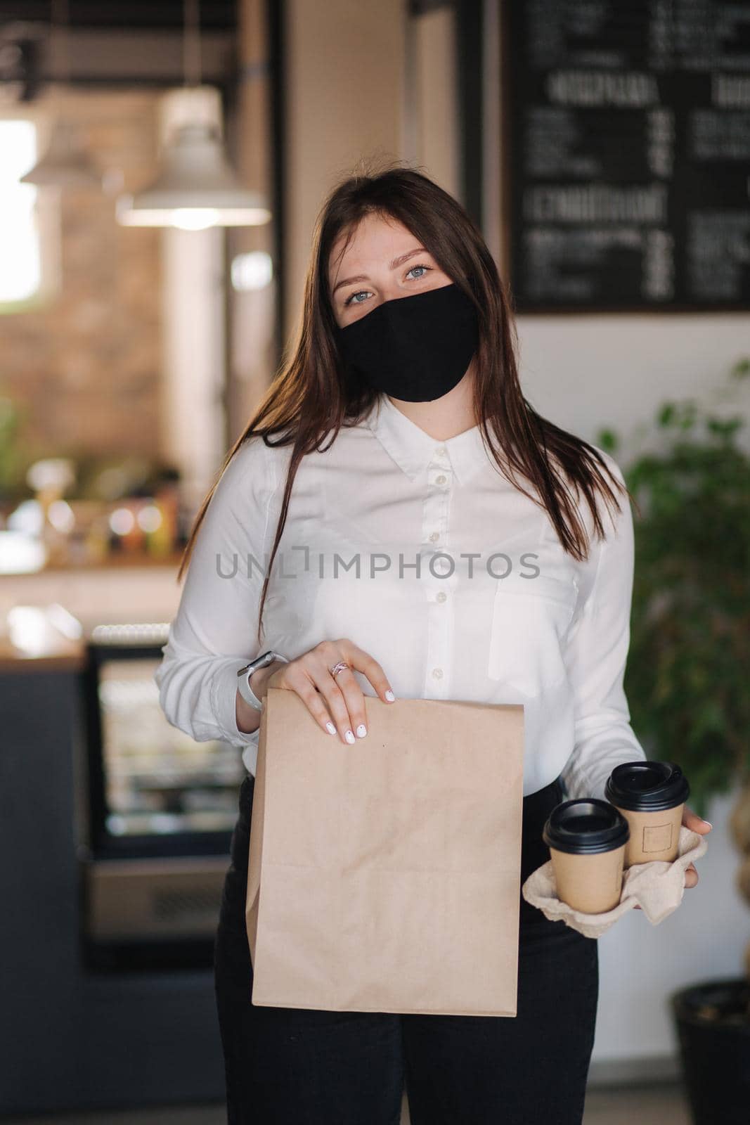 Cashier in face mask holding package with food and two cups of coffee. Contactless payments concept at cafe. Take away food during qurantine.