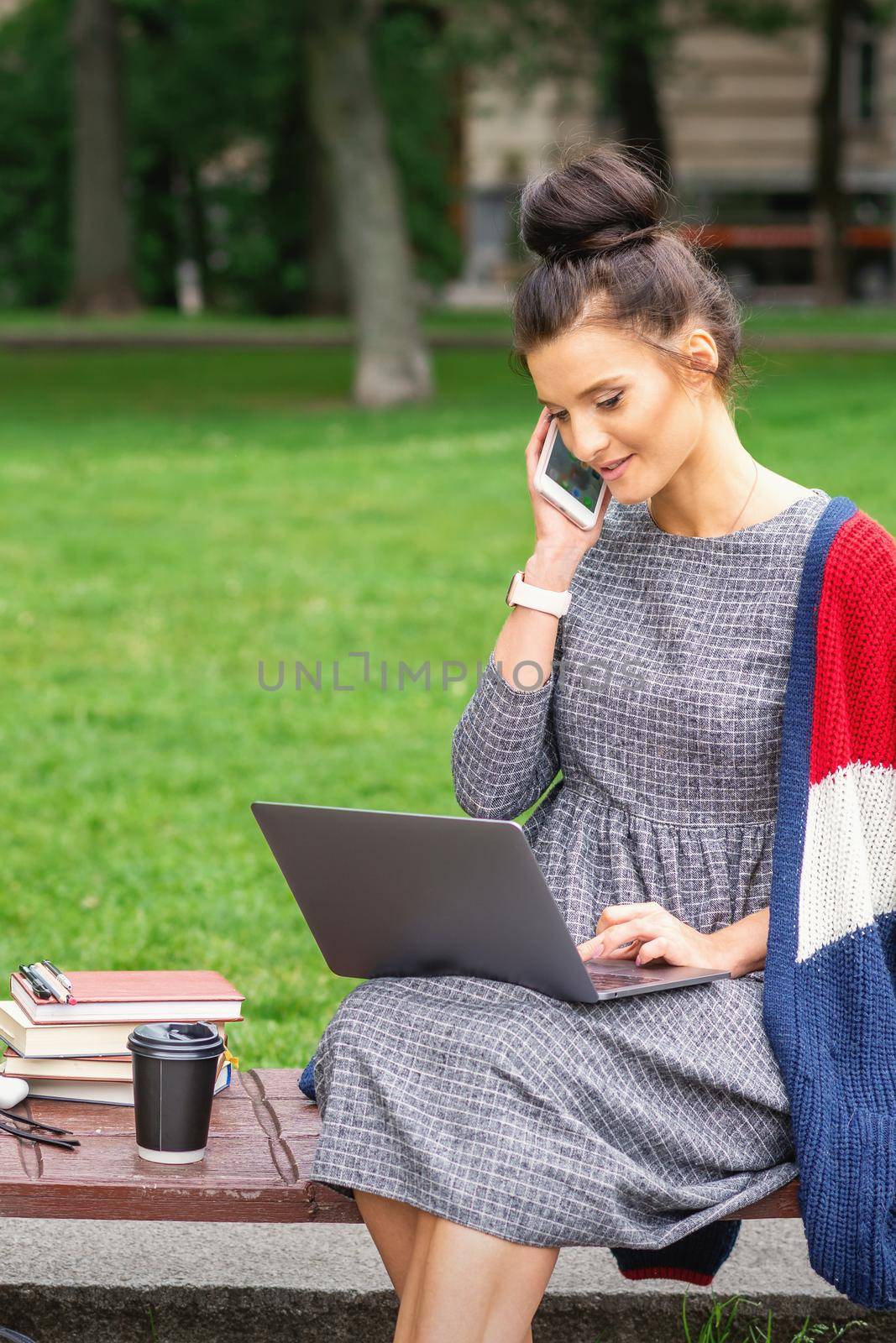Student young woman is talking by smartphone using laptop on the bench in the park