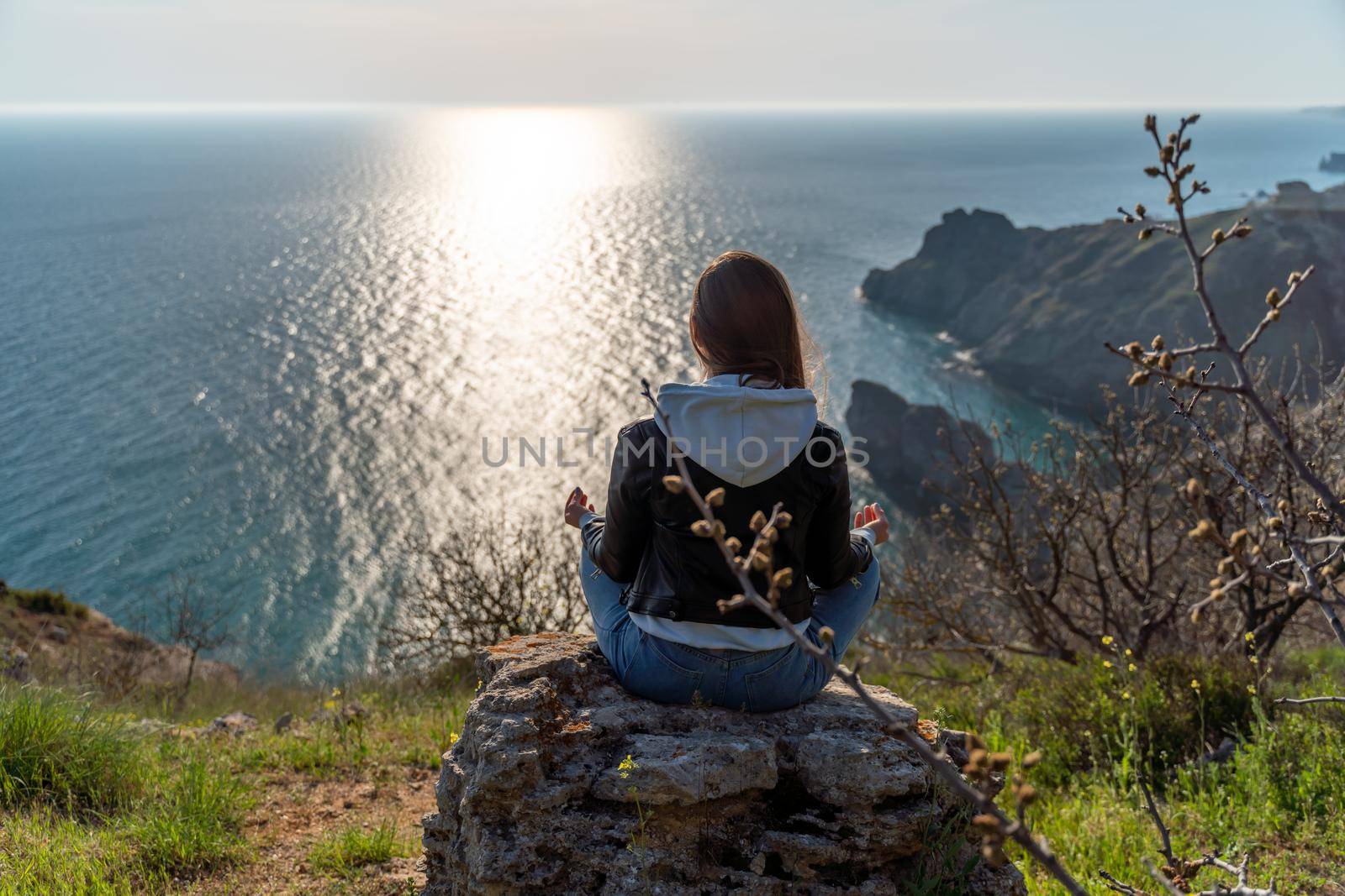 Woman tourist enjoying the sunset over the sea mountain landscape. Sits outdoors on a rock above the sea. She is wearing jeans, a blue hoodie and a black leather jacket. Healthy lifestyle, harmony and meditation.