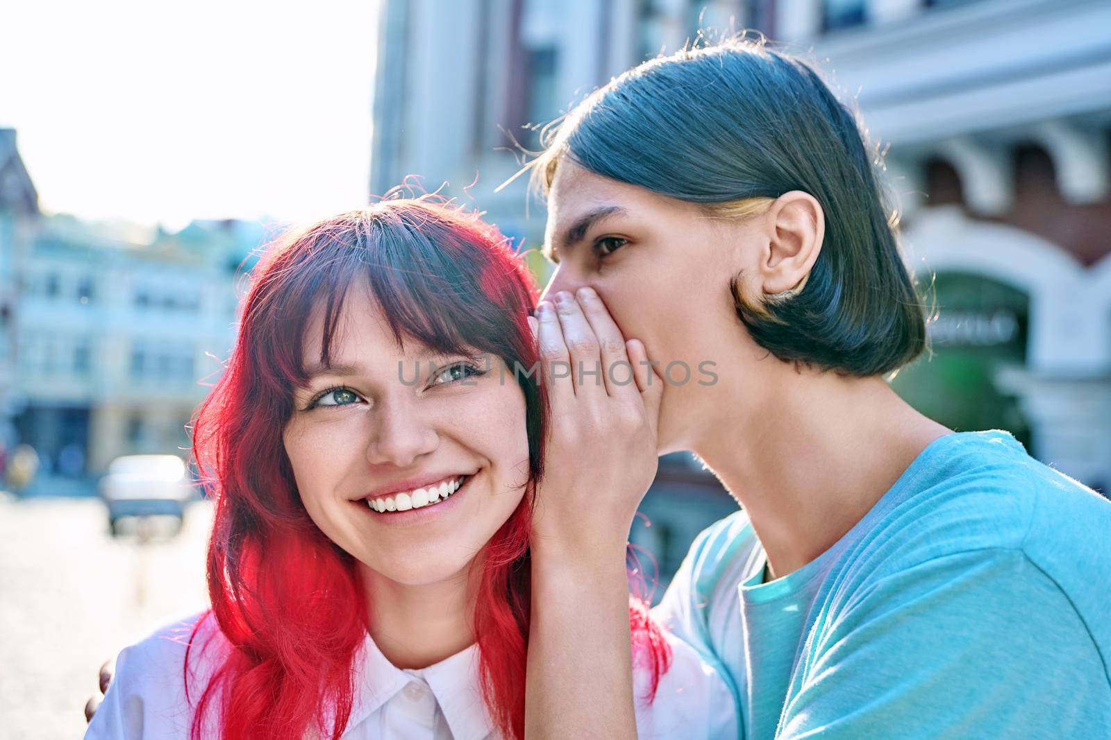 Couple of teenage students friends, guy whispers secret in ear of girlfriend on summer city street. Adolescence, friendship, relationships, surprise, emotions, youth concept