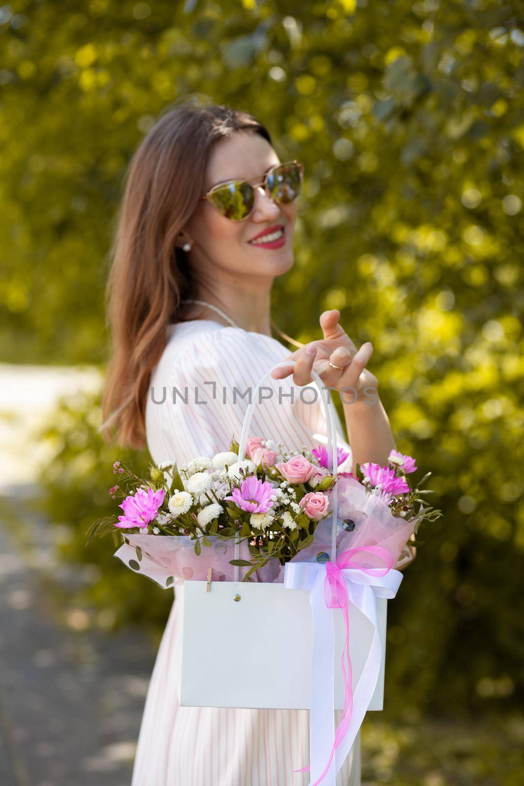 A beautiful bouquet of flowers in a box in the hands of a beautiful girl who walks along the street on a sunny day. Girl in a dress, glasses and sneakers. Focus on the background of flowers.