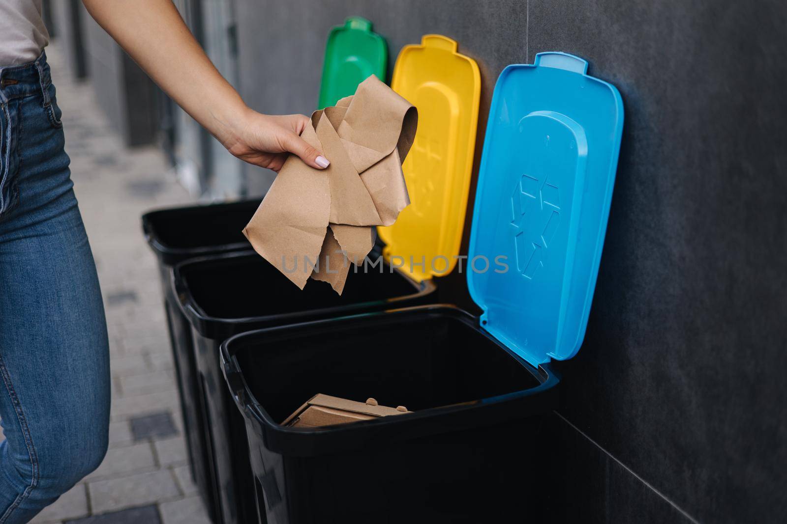 Close-up of female throwing piece of cardboard in recycling bin. Different colour of recycling bins outdoors. Side view of woman put rubbish in container.