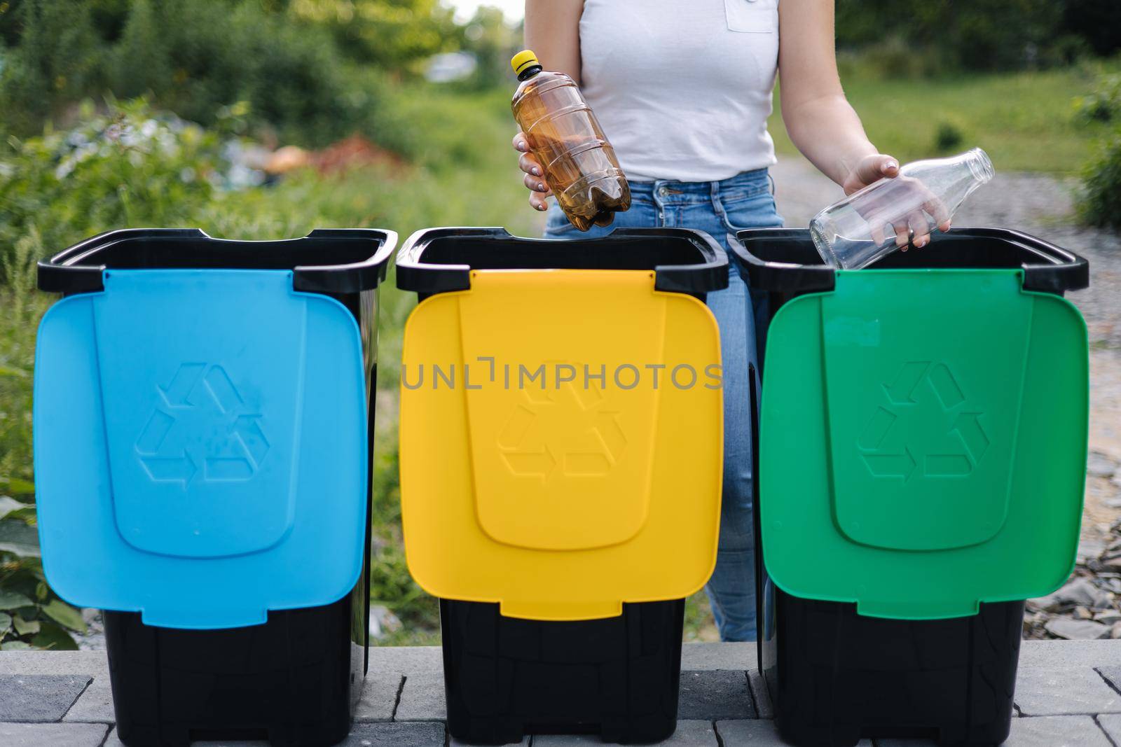 Middle selection of female hold two different bottle, plastic and glass and thowing into different recycling bins. Close-up of woman stand by blue, yeallow and green bin.