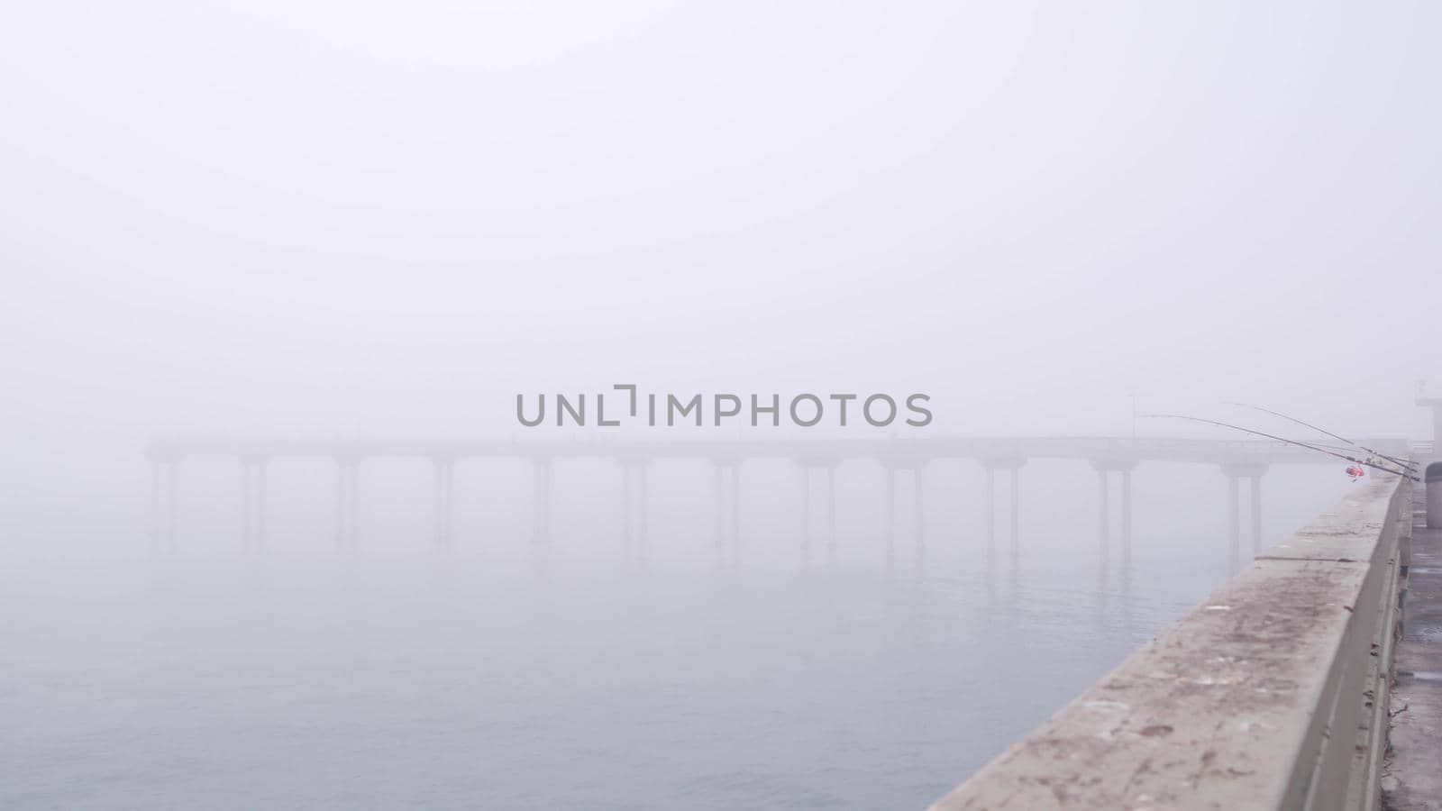 Wooden Ocean Beach pier in fog, misty California coast, USA. Foggy moody cloudy weather on San Diego shore. Calm tranquil silent atmosphere. Waterfront boardwalk. Fishing or recreational angling rod.