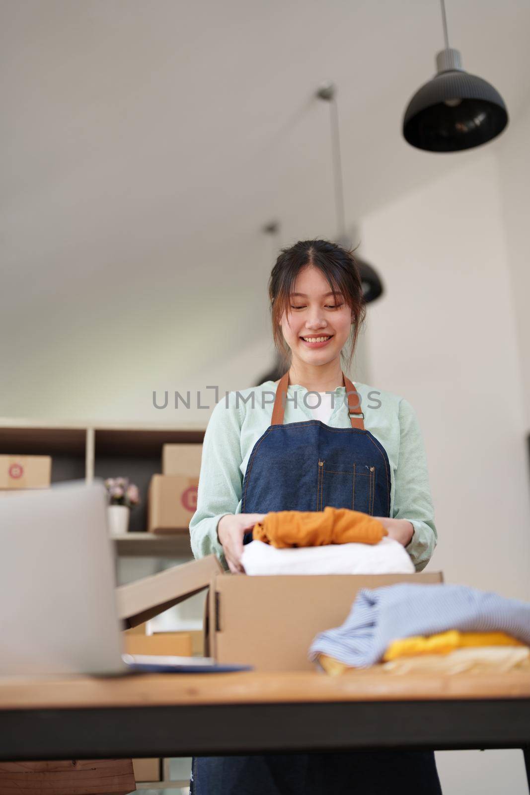 Portrait of a small startup Asian female entrepreneur SME owner picking up a yellow shirt before packing it in an inner box with a customer. Online Business Ideas and Freelance.