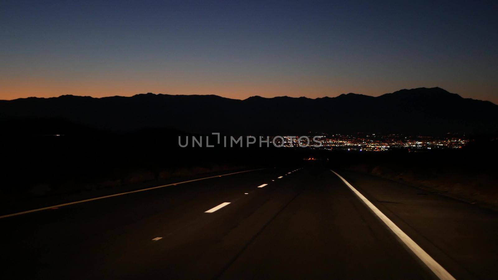 Car driving on desert road at night, freeway or highway in twilight dusk, perspective view thru windscreen or windshield. Road trip in California USA. Valley and mountains on horizon, dividing line.