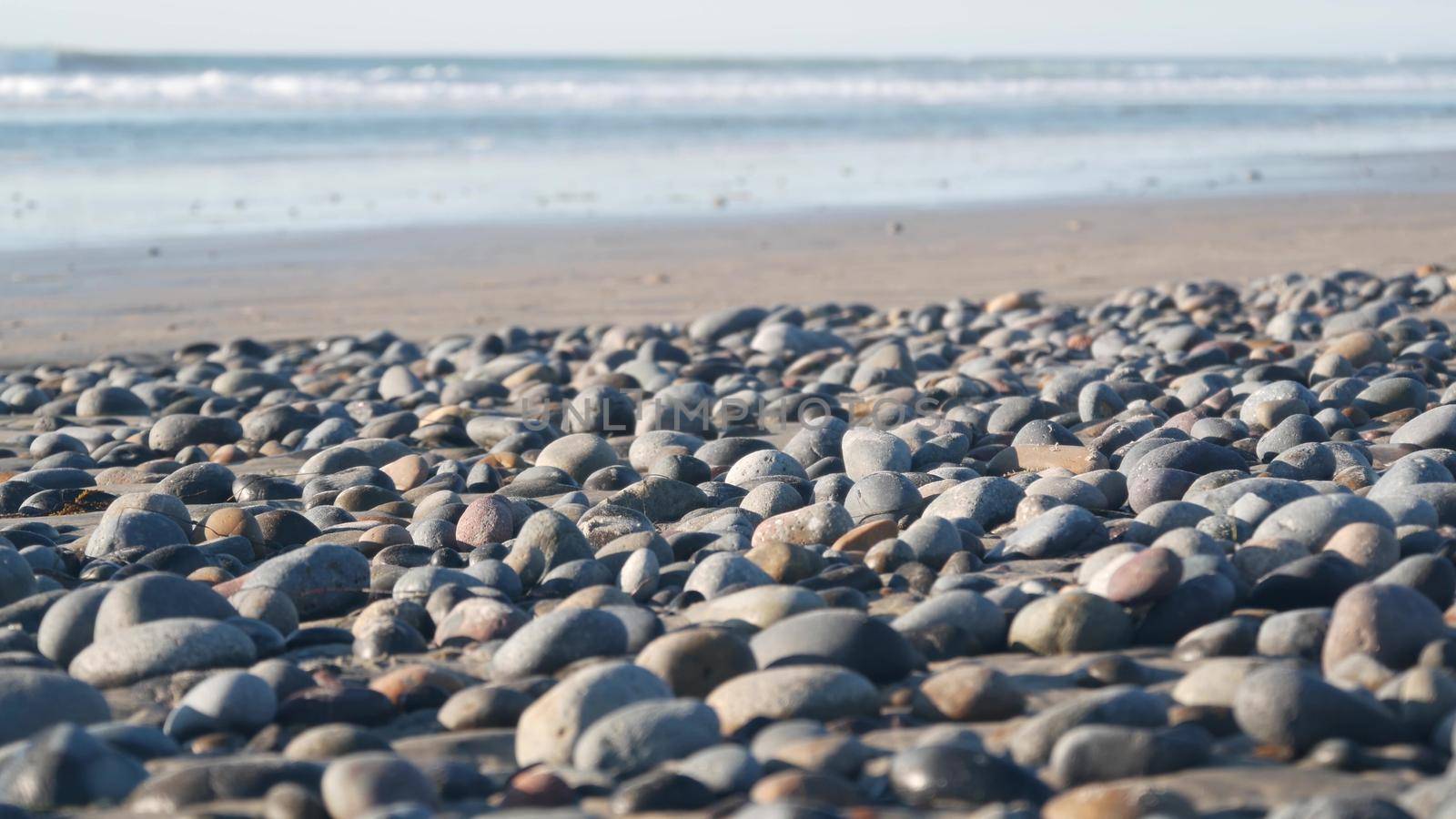 Pebble rocks on sandy beach by ocean sea water waves, round smooth stones or boulders low angle view in soft focus. California coast vacations aesthetic, USA. Seascape natural background with bokeh.
