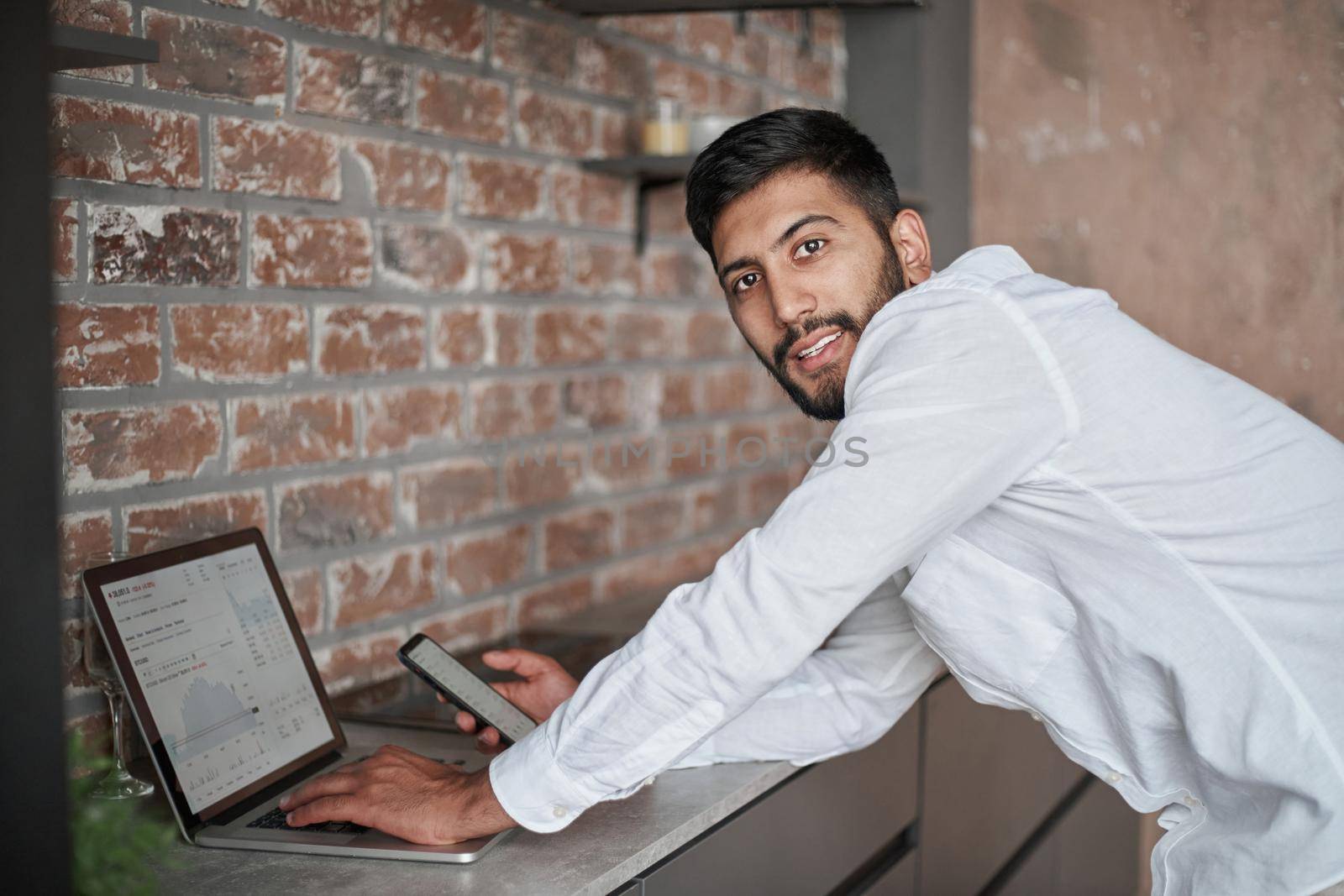 young man using a laptop and a smartphone standing in the kitchen. by SmartPhotoLab