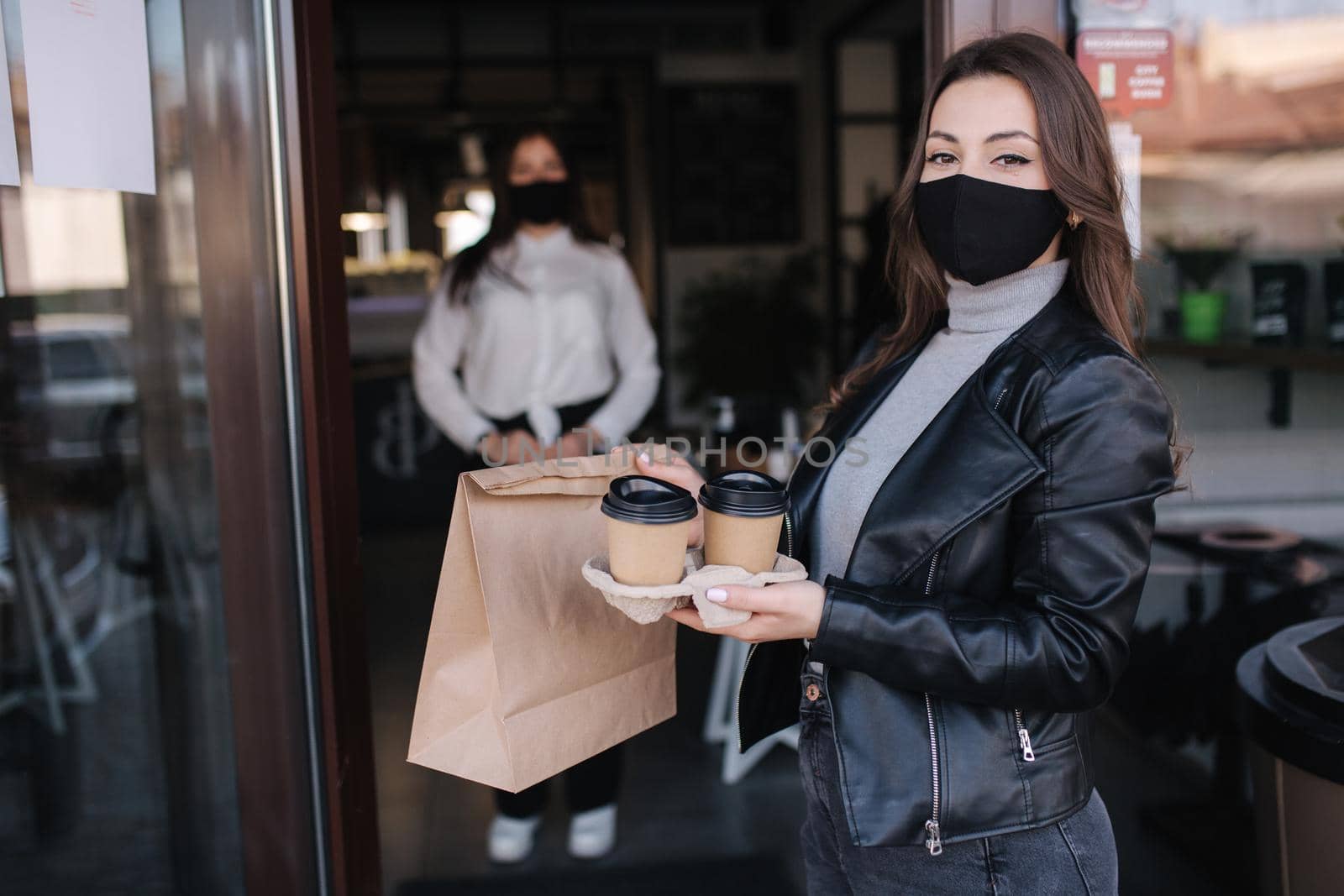 Attractive female customer in face mask standing outdoors by cafe and take away food and coffee. Background of cashier in mask waiting for next customer. New norm during quarantine.