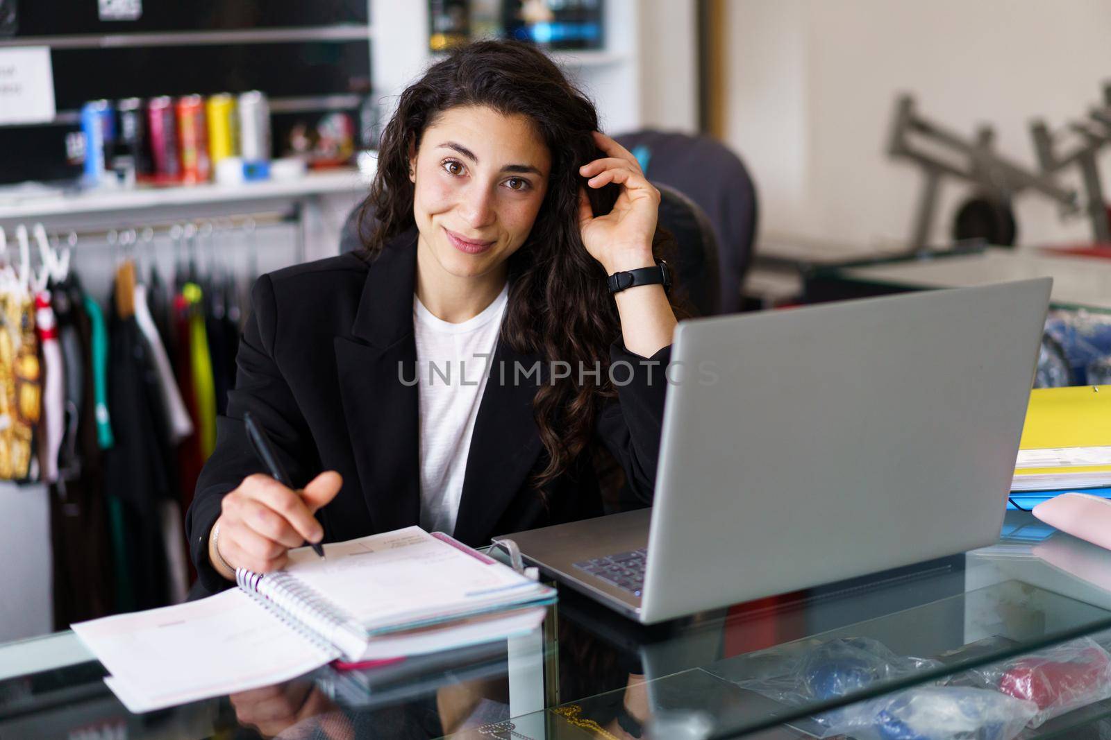 Positive young woman, in smart casual clothes, looking at camera with smile and touching dark hair, while sitting at desk and making notes during psychology session in gym