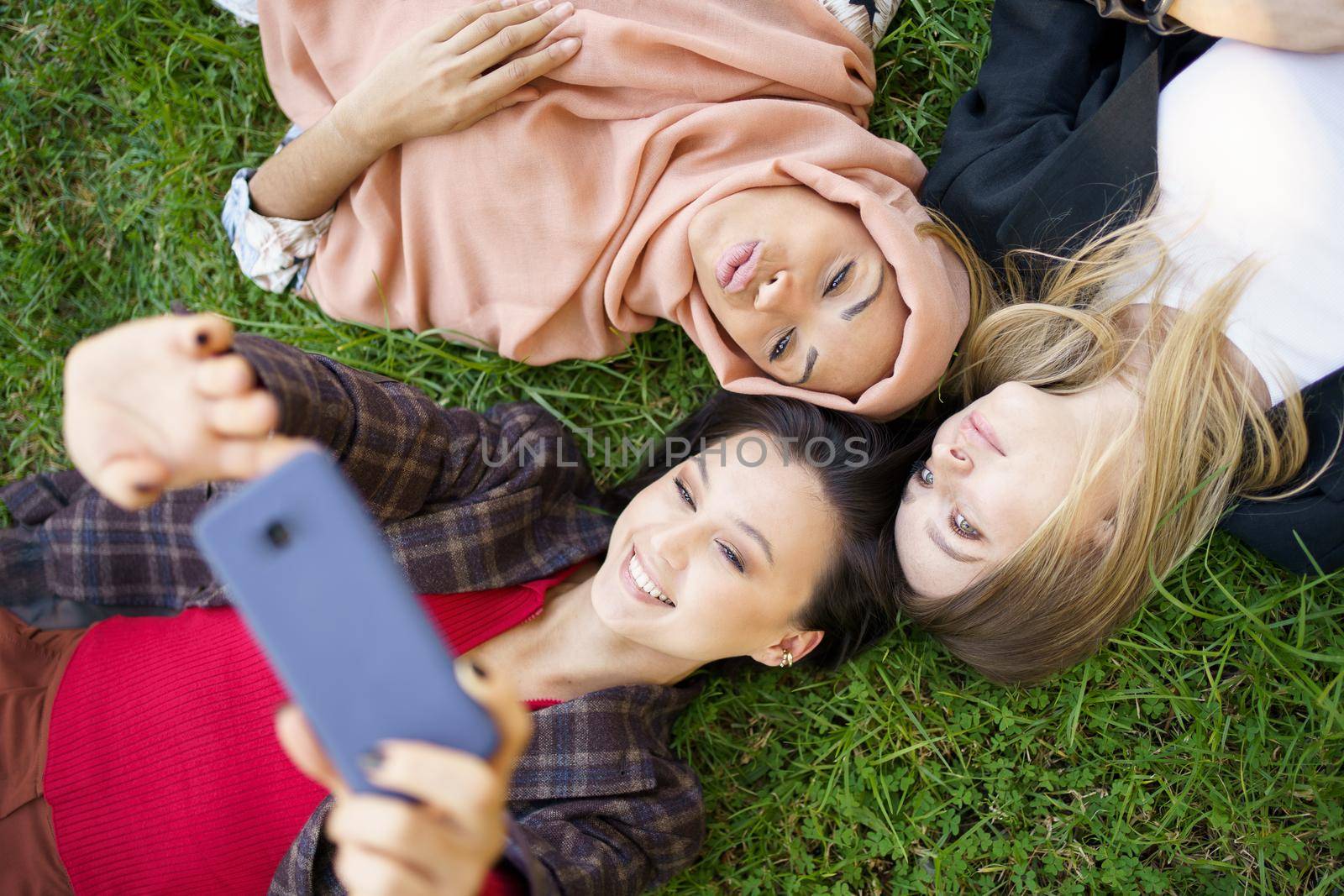 Top view of multiethnic young ladies lying on grass and taking self portrait on cellphone