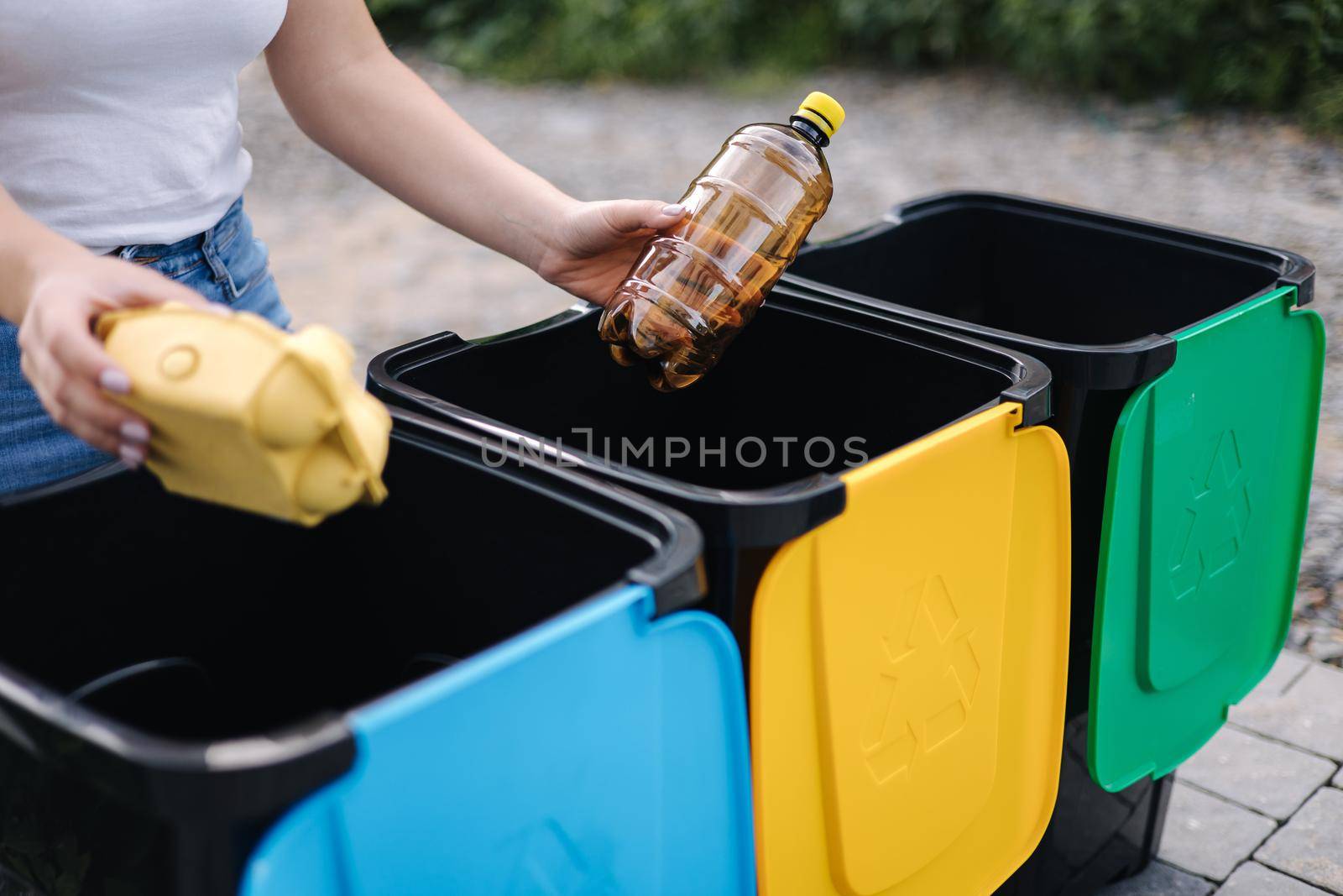 Middle selection of female hold two different type of rubbish, plastic bottle and egg carton and thowing into different recycling bins. Close-up of woman stand by blue, yeallow and green bin.