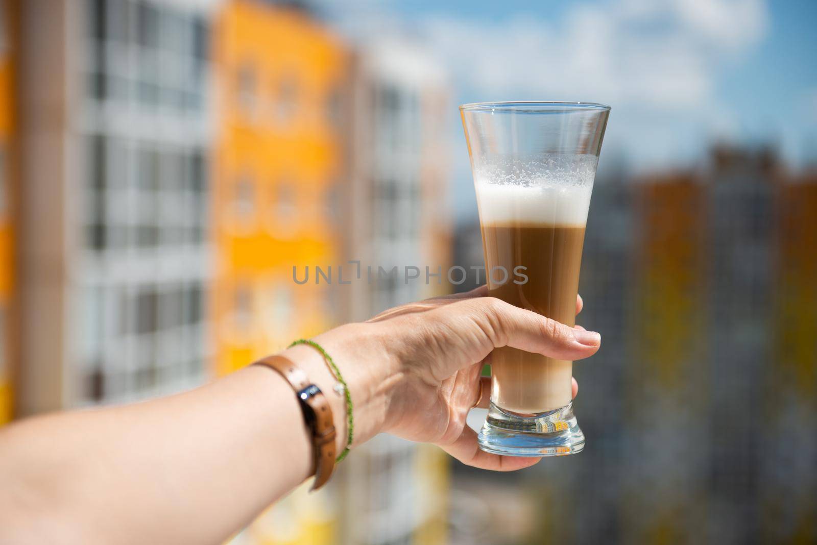 multilayer coffee or cappuccino in a glass on a woman's hand stretched out into the window against the background of an apartment building