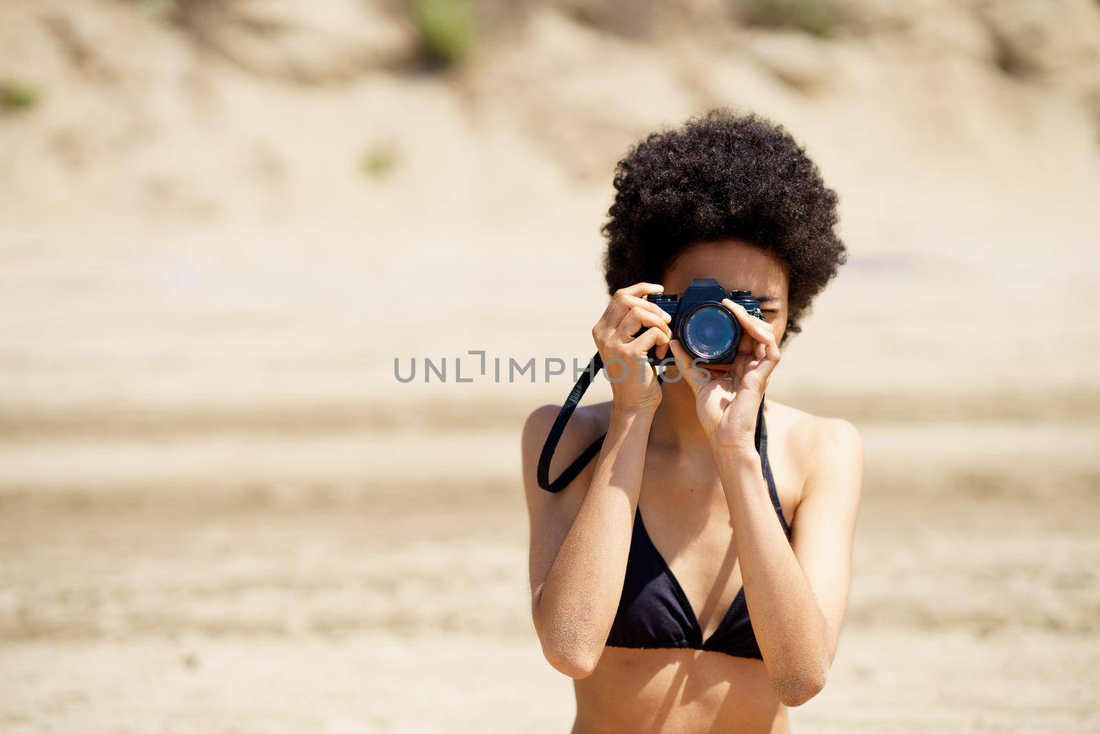 African American female photographer in swimwear taking picture on photo camera while standing on sandy shore during trip in tropical resort