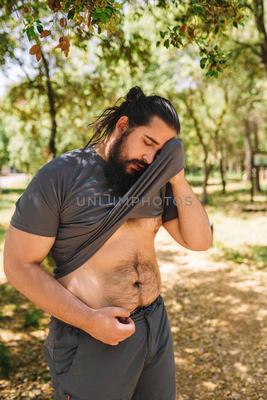 young man practising sport outdoors wiping sweat from his forehead with his t-shirt. athlete preparing for a marathon. health and wellness lifestyle. by CatPhotography