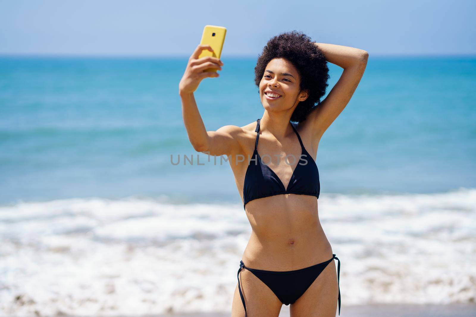 Optimistic fit African American female traveler in swimwear taking self portrait on smartphone with hand behind head while standing near waving sea in summer