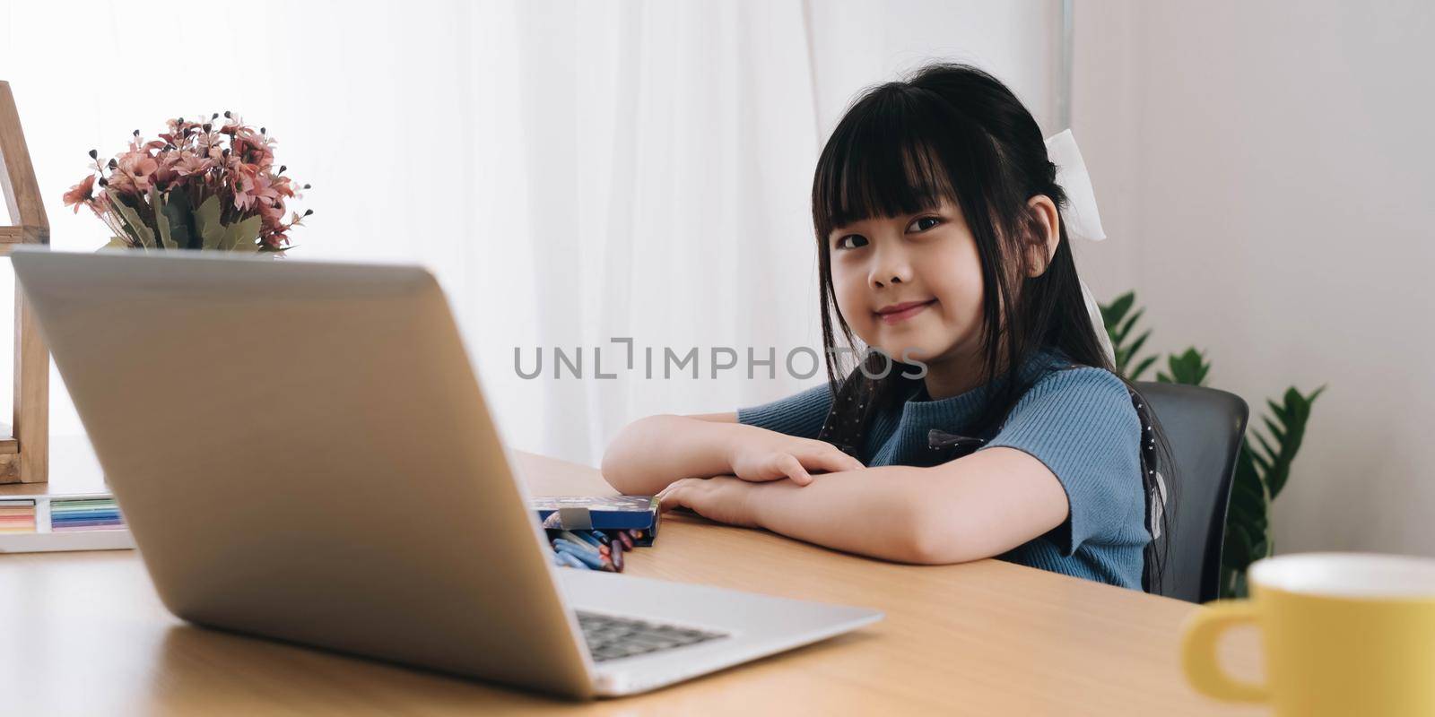 smiling little girl looking at the camera sitting at a table studying at home cute kid doing homework holding a pen taking notes watching webinars.