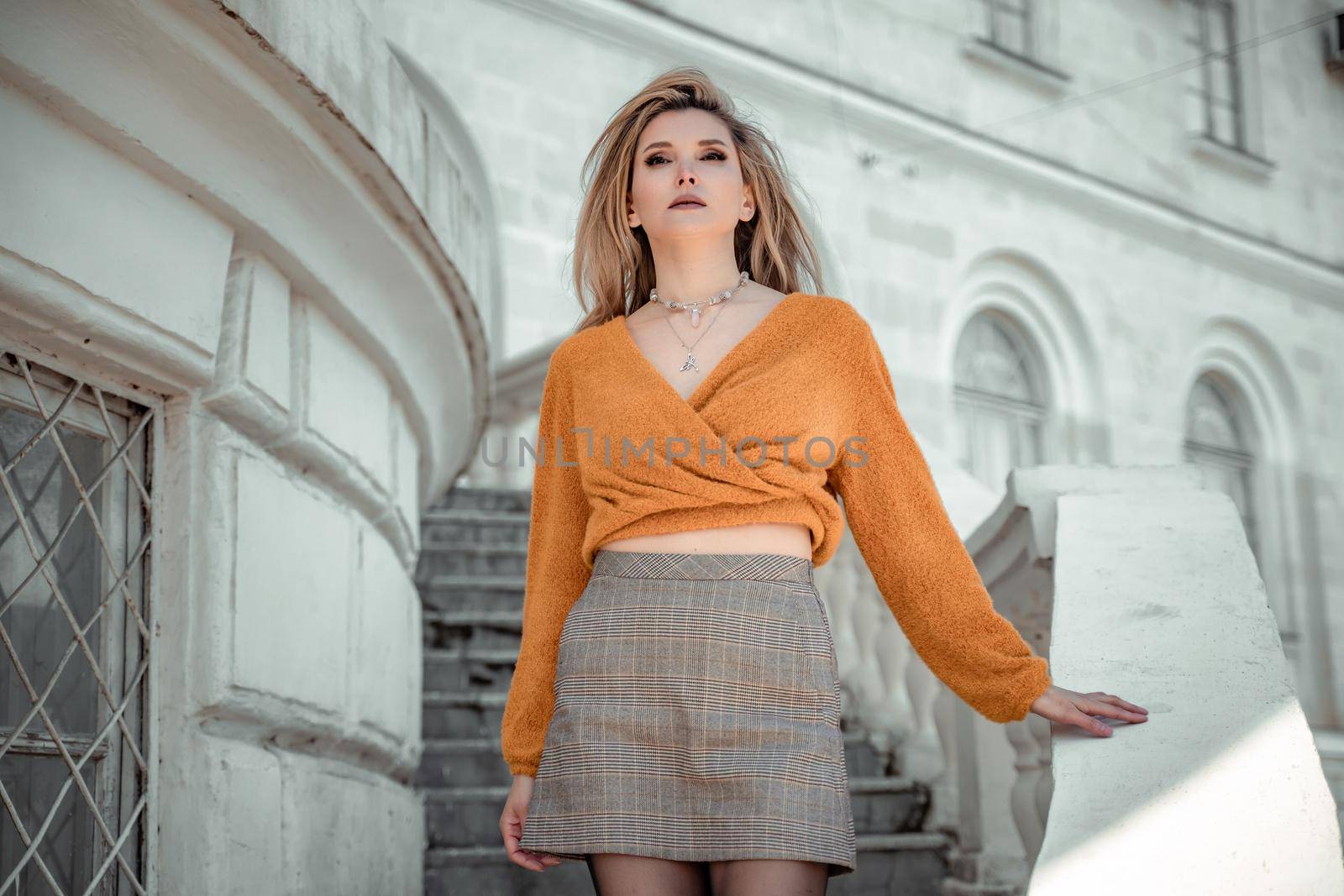 A middle-aged woman looks like a good blonde with curly beautiful hair and makeup on the background of the building. She is wearing a yellow sweater. by Matiunina