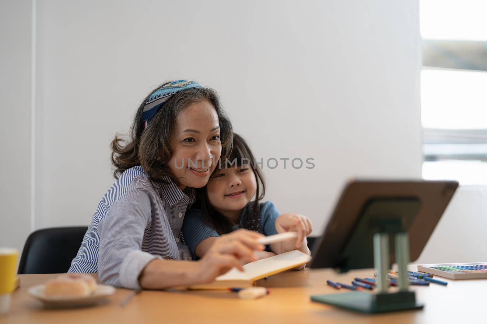 Loving senior grandmother hug shoulders of small girl grandchild assist in preparing homework support give advice. Friendly aged woman teacher watching little kid pupil writing maths problem solution.