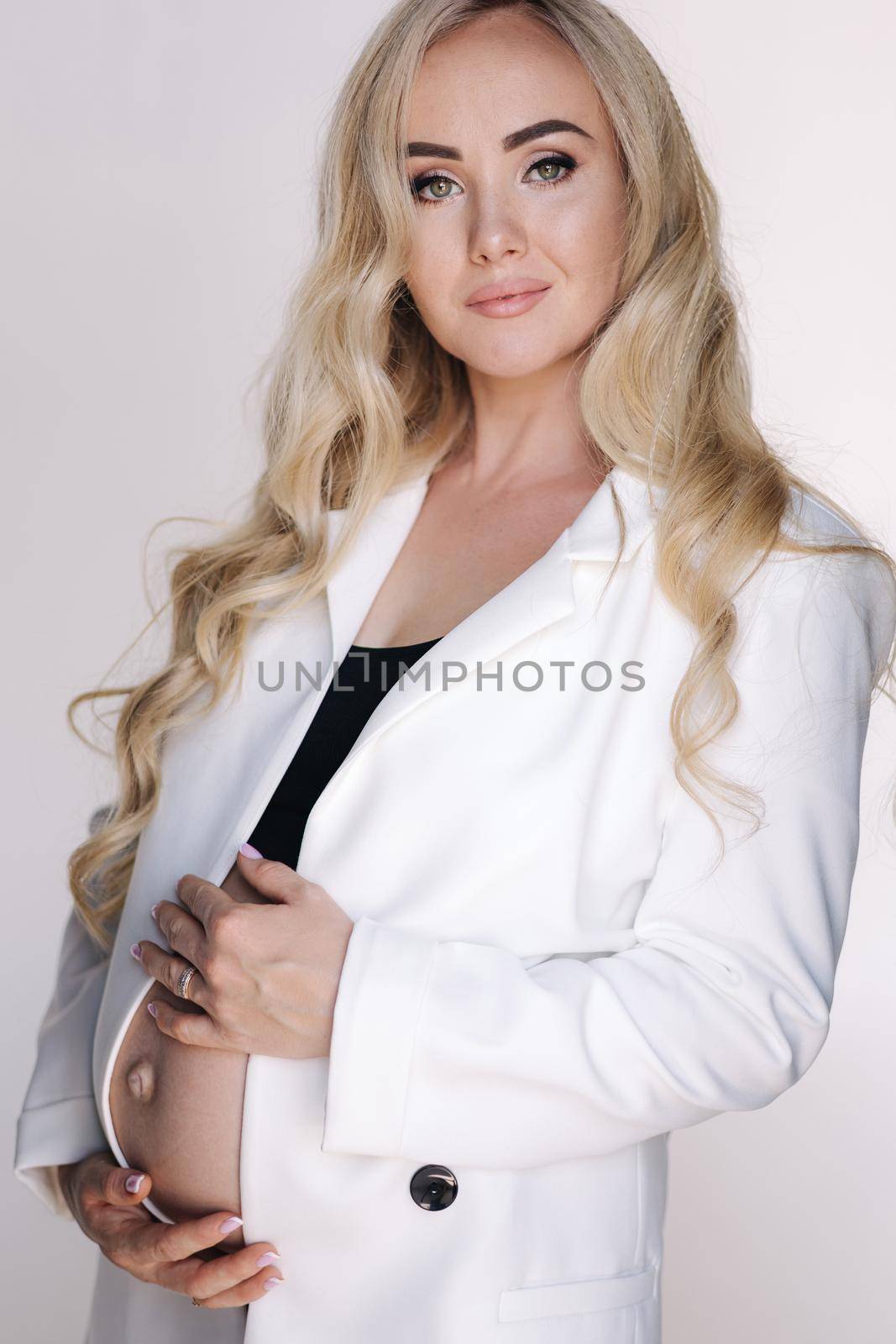Portrait of pregnant woman in studio on white background. Beautiful future mother put her her hands on her belly. Blond hair woman in white jaket.