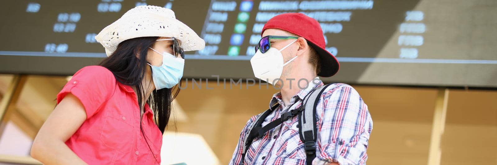 Low angle of couple waiting for flight holding tickets and wearing face masks. Man and woman going on vacation, suitcase packed. Trip, traveling concept