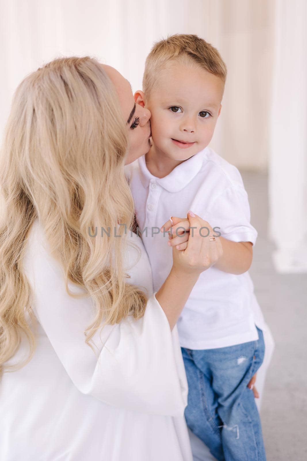 Beautiful pregnant woman with her son. Lady in elegant white dress posing to photographer in studio. Background of white tulle.