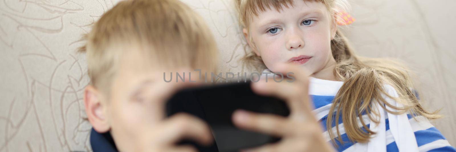 Portrait of brother and sister at home on sofa watching on smartphone screen. Kids use modern technology from young age. Addict, holiday, siblings concept