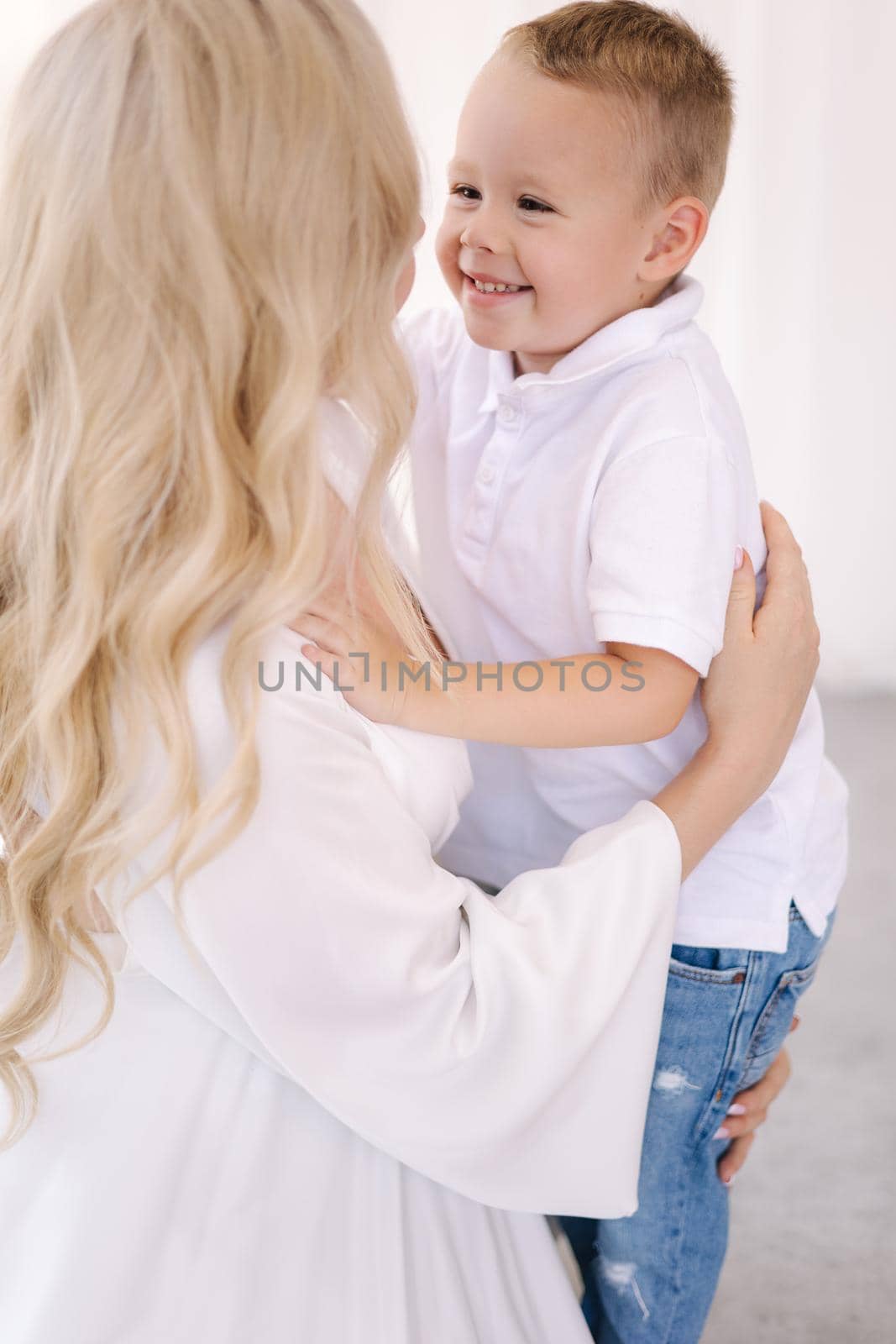 Beautiful pregnant woman with her son. Boy hug his mom and rejoices. Lady in elegant white dress posing to photographer in studio. Background of white tulle.