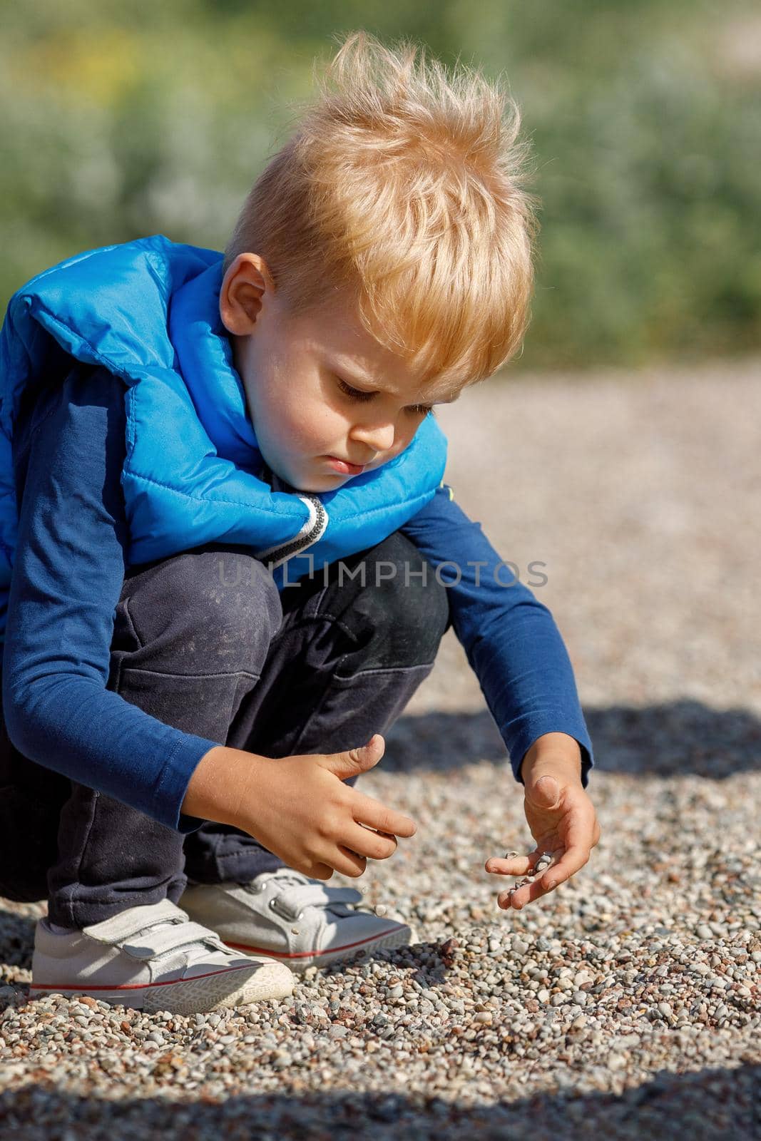 A little boy with a blue vest plays outdoors with small pebbles. The child explores the ground with his own hands, he is very interested in small pebbles.