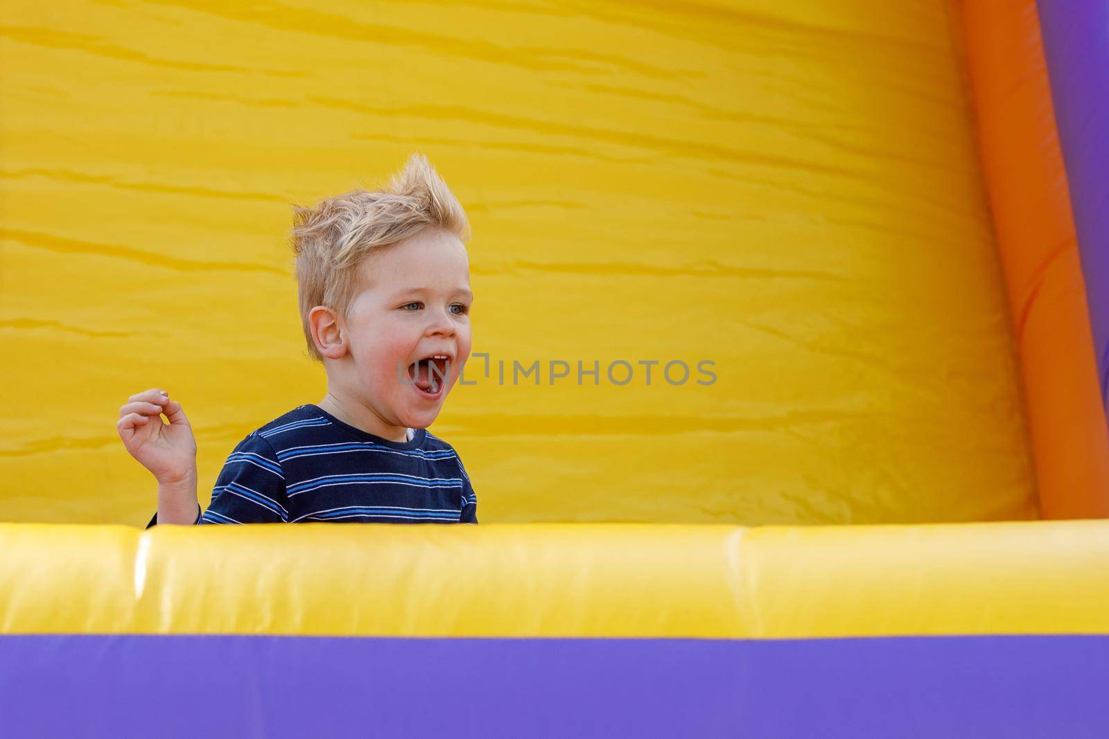 Little boy having fun in inflatable castle playground. Furious child shouts loudly. Bright yellow rubber trampoline background, there is free copy space.