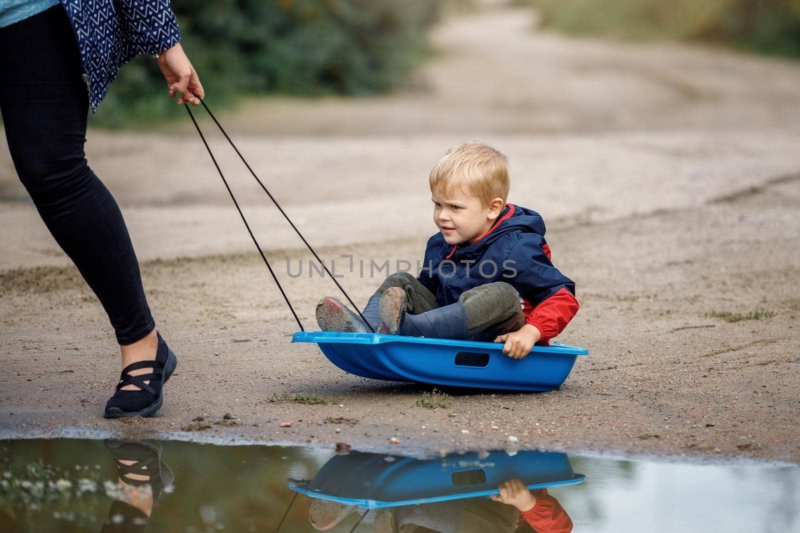 A little boy sitting on a sled skates in the mud near the swamp. Summer time after the rain, sled towing his mom they joke. by Lincikas