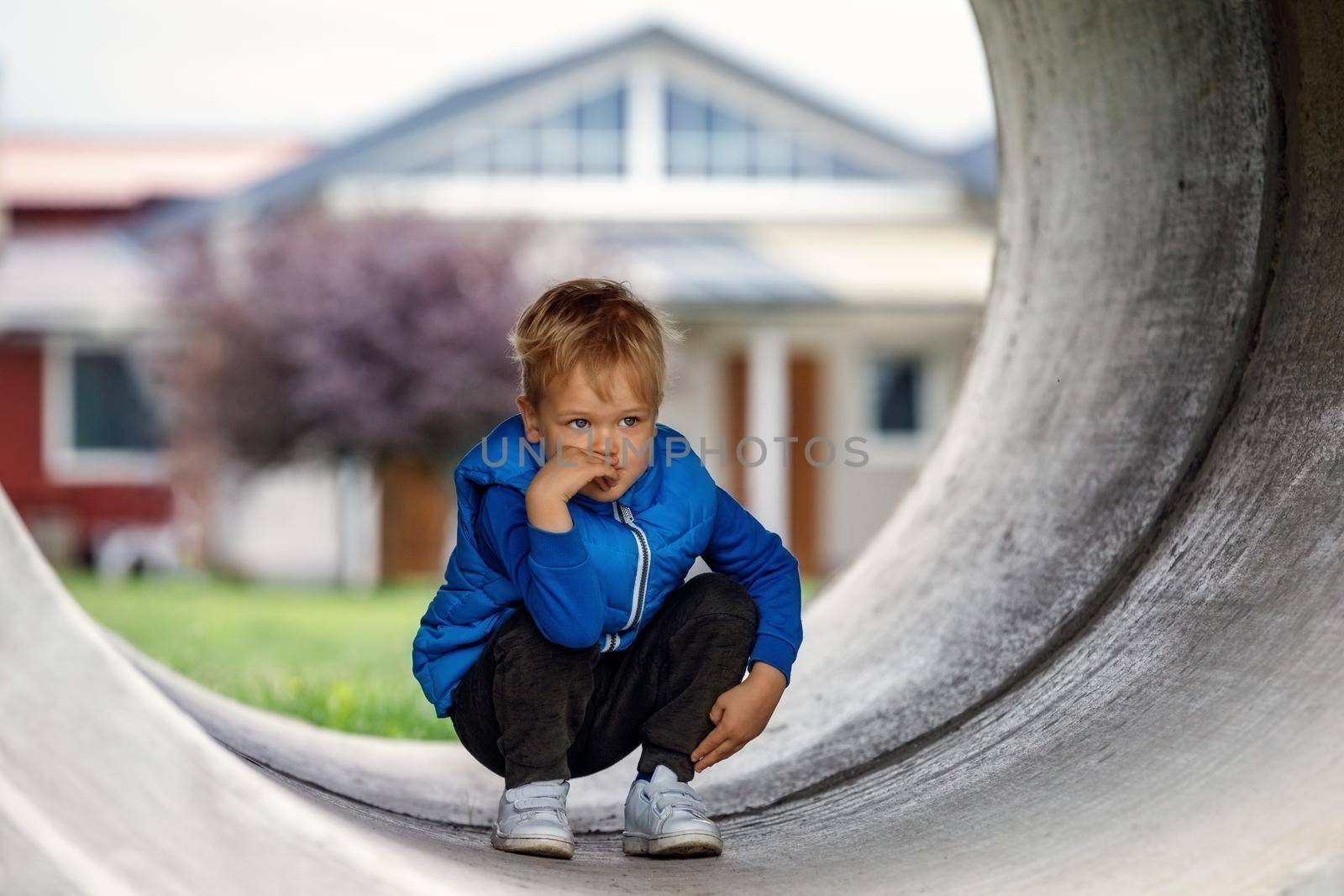 A cute kid wearing a blue vest squats in a concrete tunnel and thinks of something. Horizontal photo.