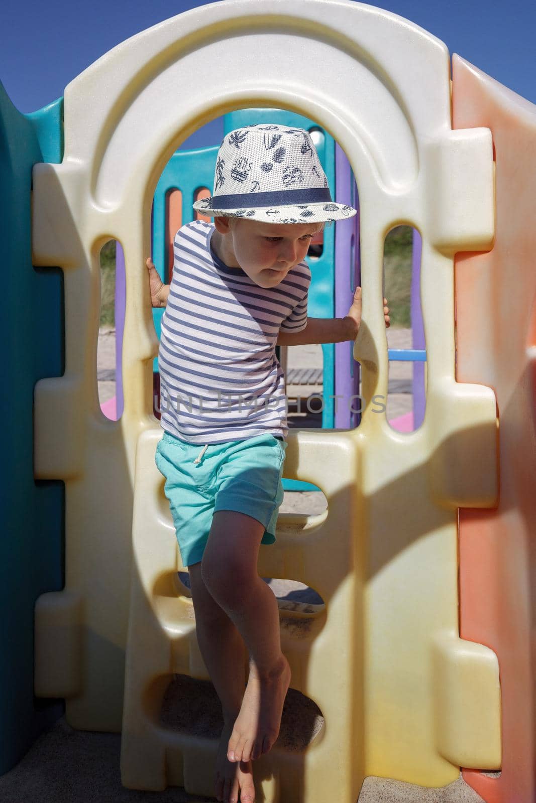 A little boy in a striped shirt performs workout exercises on a coloruful beach playground. by Lincikas