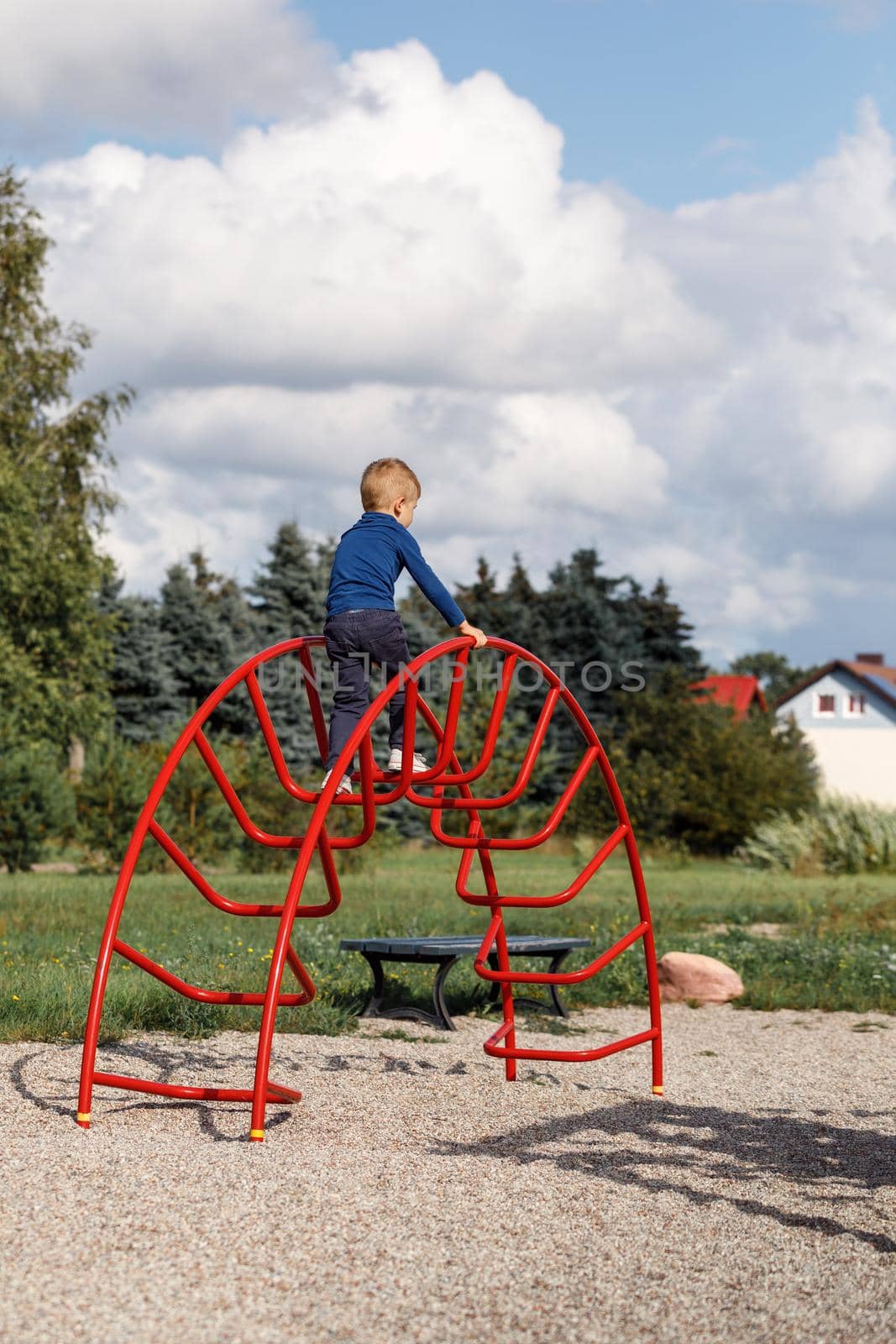A little very brave boy climbs a metal, big red arched ladder on the playground. The concept of parental care and safety in children's play.