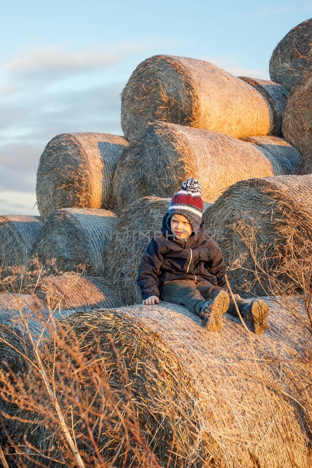 A cute little boy in autumn clothes and a knitted hat poses against a background of golden hay bales. Vertical photo.