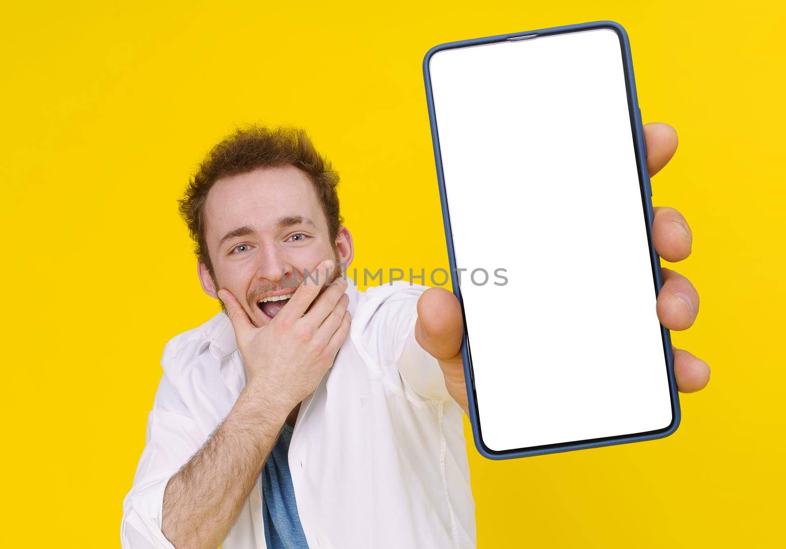 Cant believe, shocked young man with smartphone showing a white empty screen isolated over yellow game, bet, lottery win. Product placement. Mobile App Advertisement.