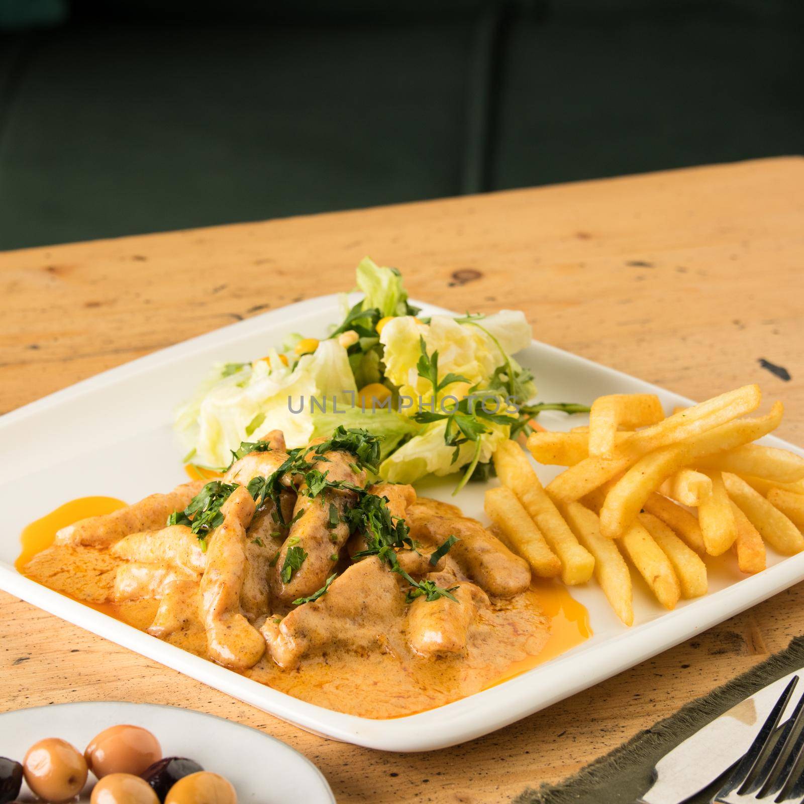 A closeup of a meal with chicken, french fries, salad and olives on a table