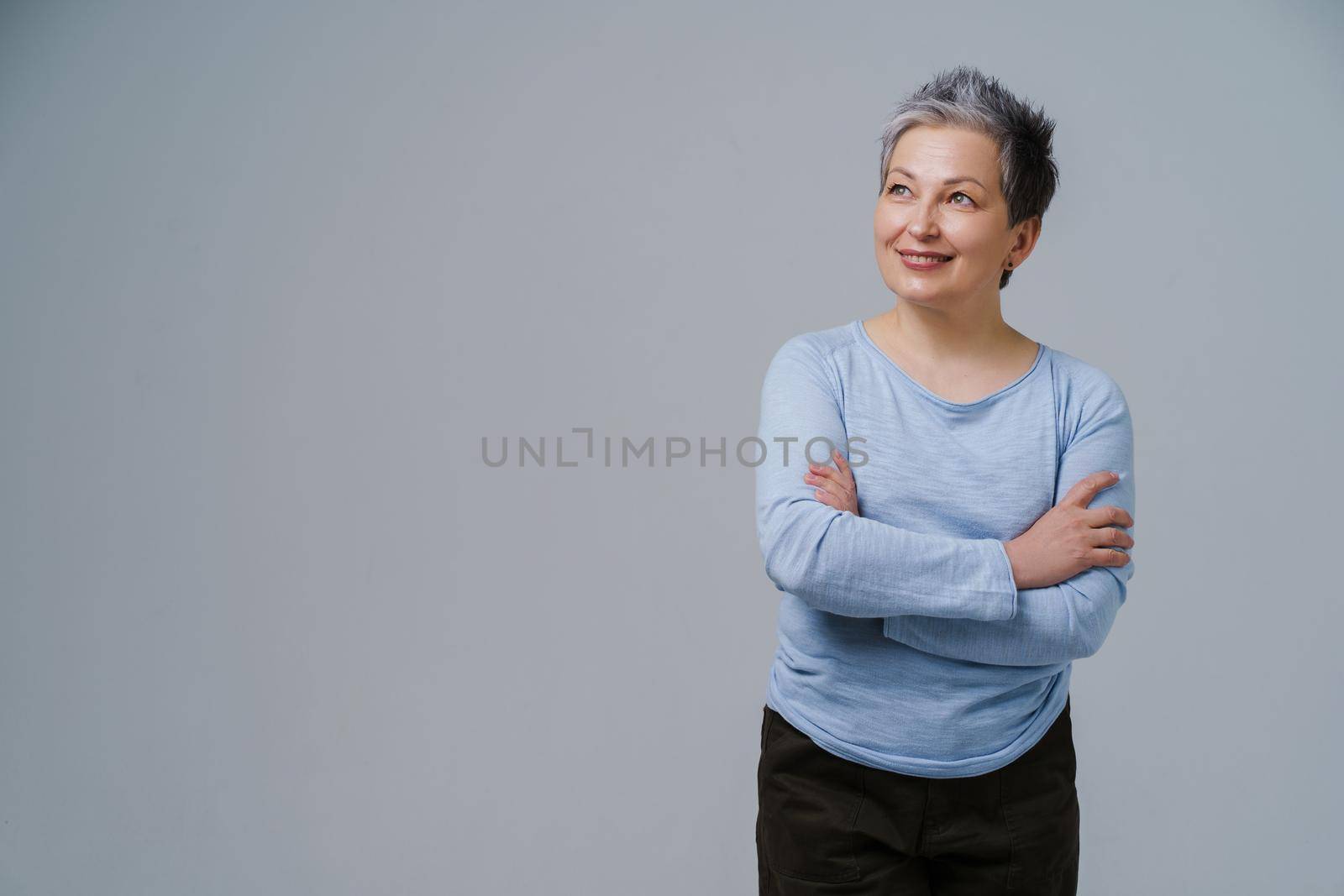 Mature grey hair woman posing with hands folded in blue blouse looking sideways up, copy space on left isolated on white background. Healthcare concept. Aged beauty concept. Copy space.