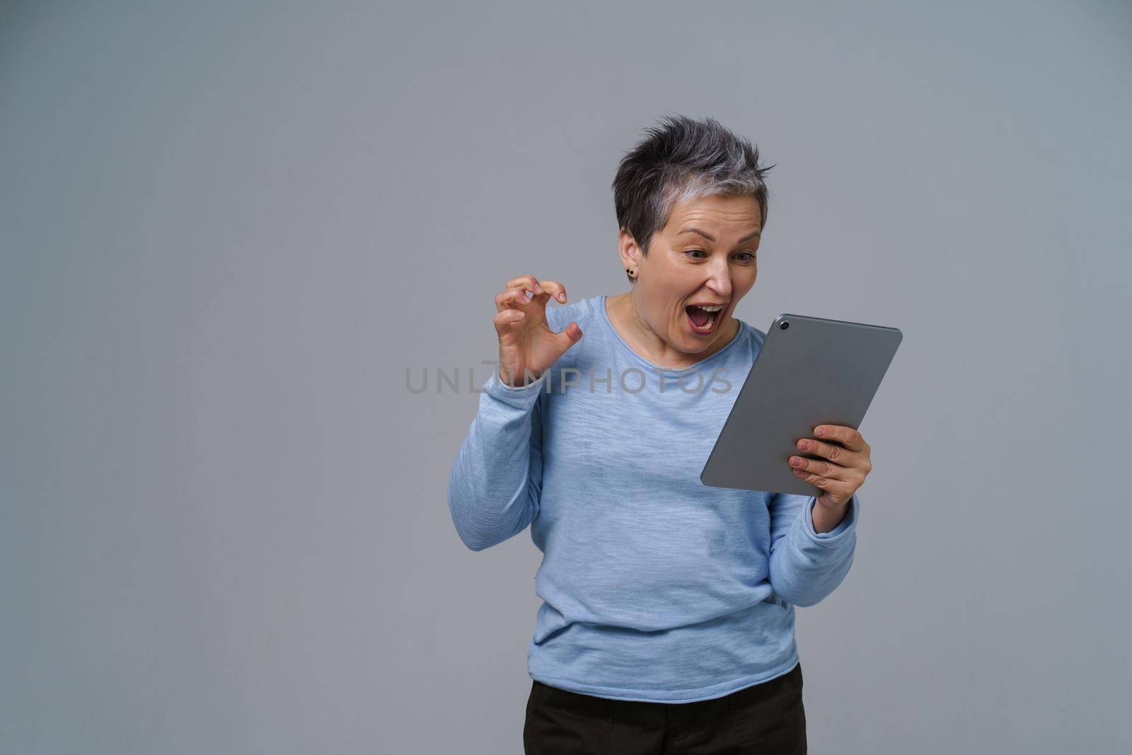 Inspired, shocked mature grey hair businesswoman with digital tablet in hand working, shopping, gaming online. Pretty woman in 50s in blue blouse isolated on white.