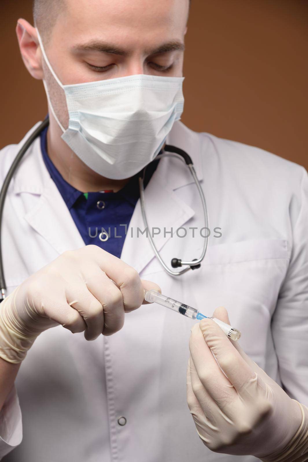 Studio portrait of a male doctor in a white coat with a syringe and an ampoule of a vaccine in his hands. Caucasian doctor on brown background by yanik88