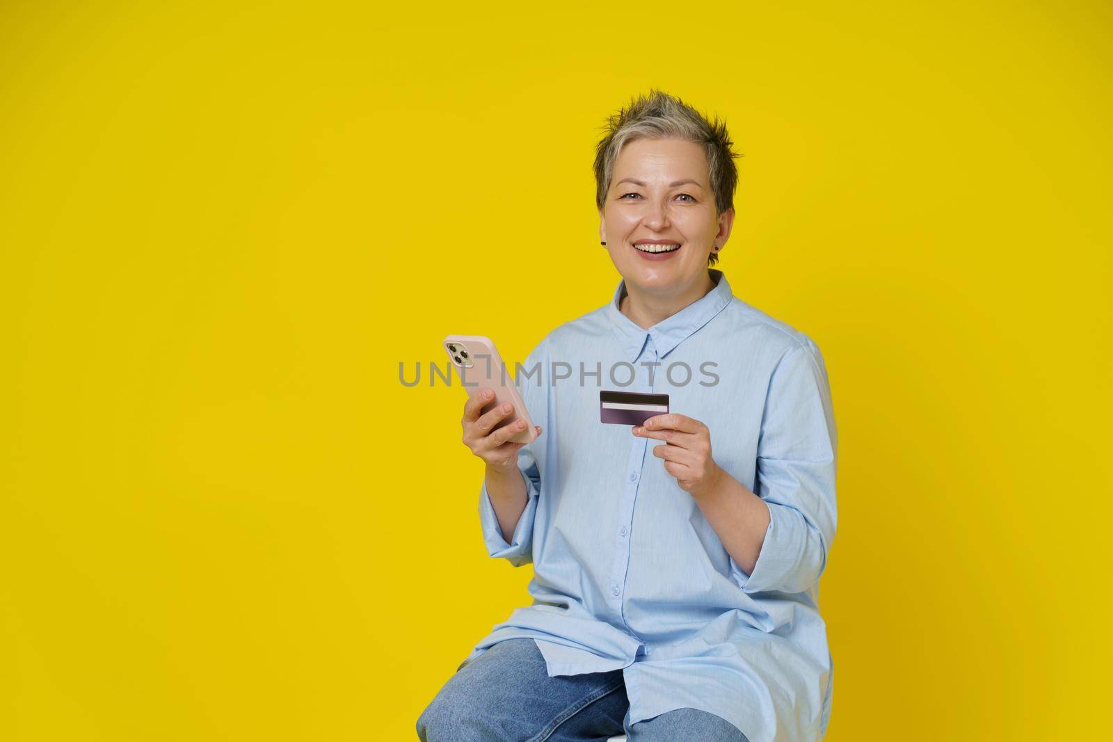 Grey haired woman hold phone and debit or credit card in hand making online payment or shopping. Pretty mature woman in blue shirt and jeans isolated on yellow background by LipikStockMedia