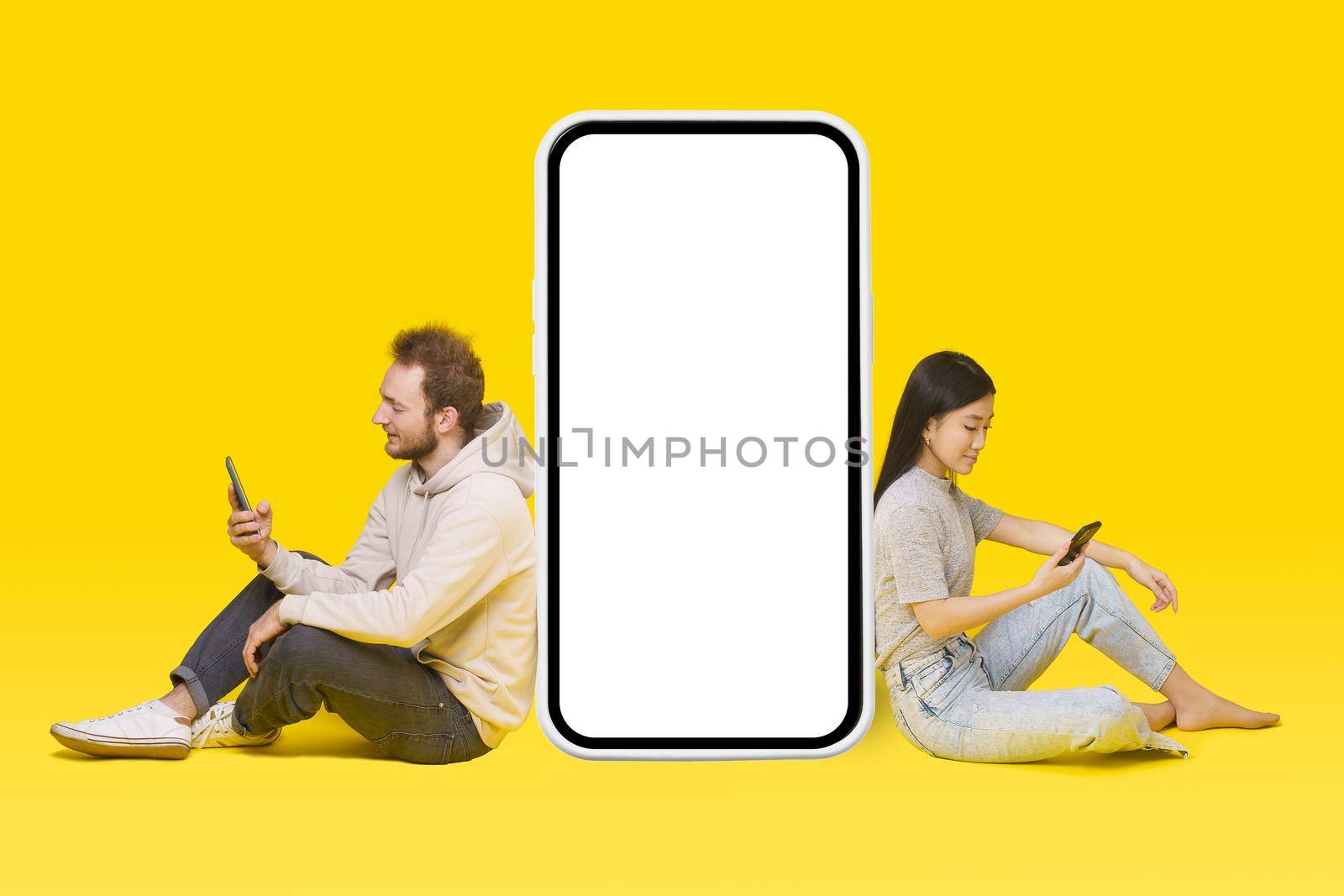Caucasian guy and asian girl sitting on floor with phones in hands leaned on huge, giant smartphone blank white screen, mobile app advertisement isolated on yellow. Product placement.