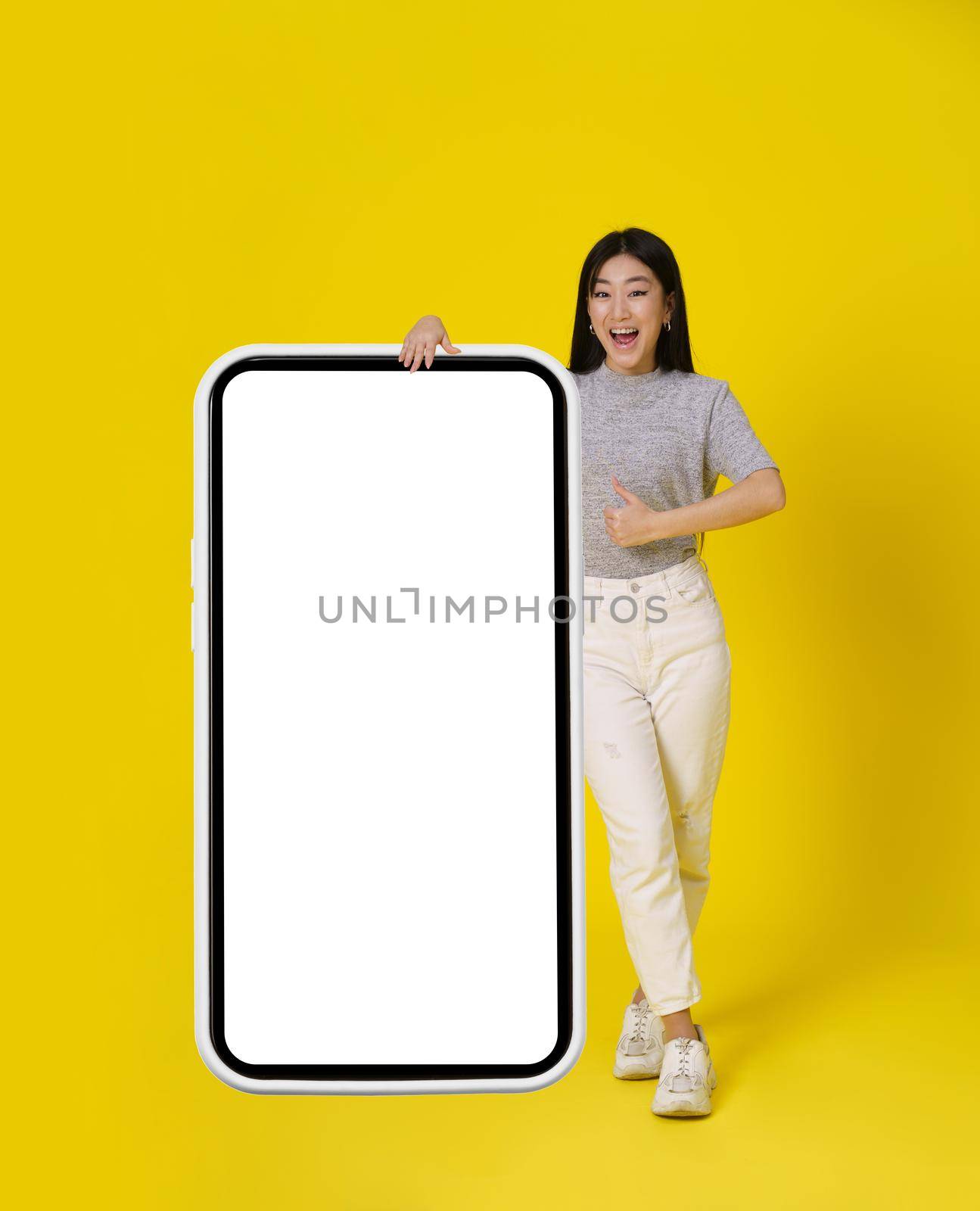 Charming asian girl gesturing thumb up stand near huge, giant smartphone with white screen happy dressed in casual isolated on yellow background. Free space mock up. Product placement mobile app add by LipikStockMedia