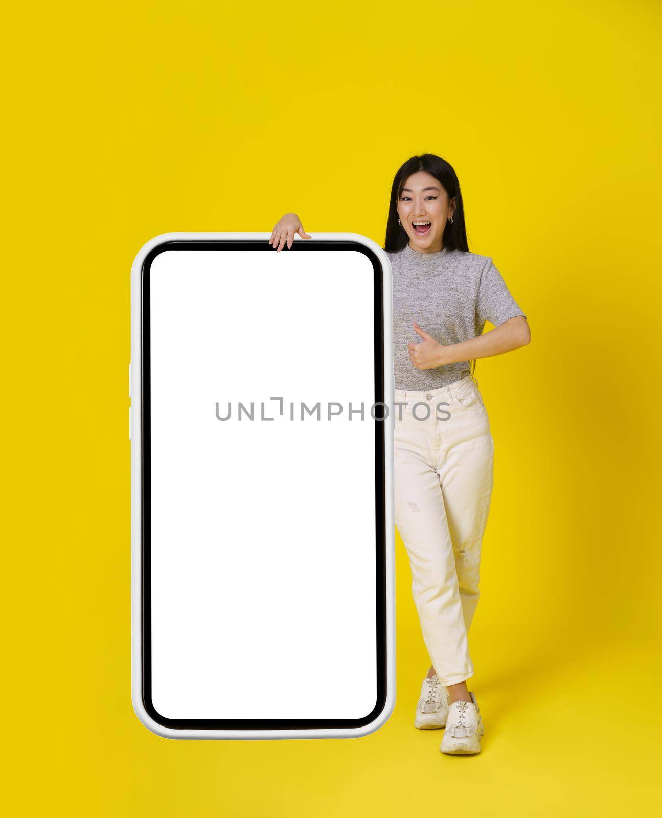 Charming asian girl gesturing thumb up stand near huge, giant smartphone with white screen happy dressed in casual isolated on yellow background. Free space mock up. Product placement mobile app add.